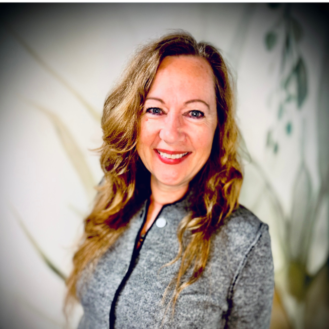A woman with curly brown hair, wearing glasses, smiling, standing in front of a bookshelf and some green plants.