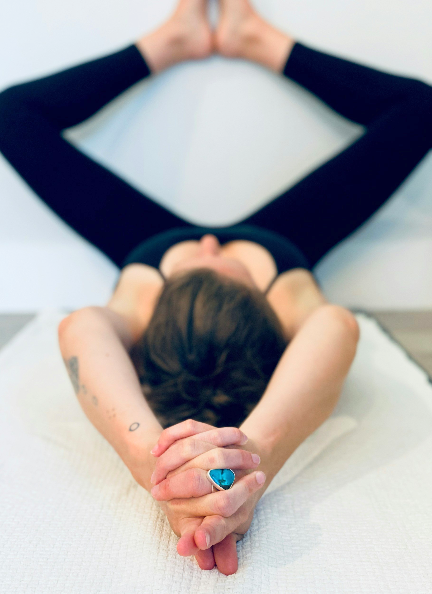 Person lying on a white surface with hands clasped behind their head, wearing a black top, with tattoos on their left arm and a large turquoise ring on their finger.