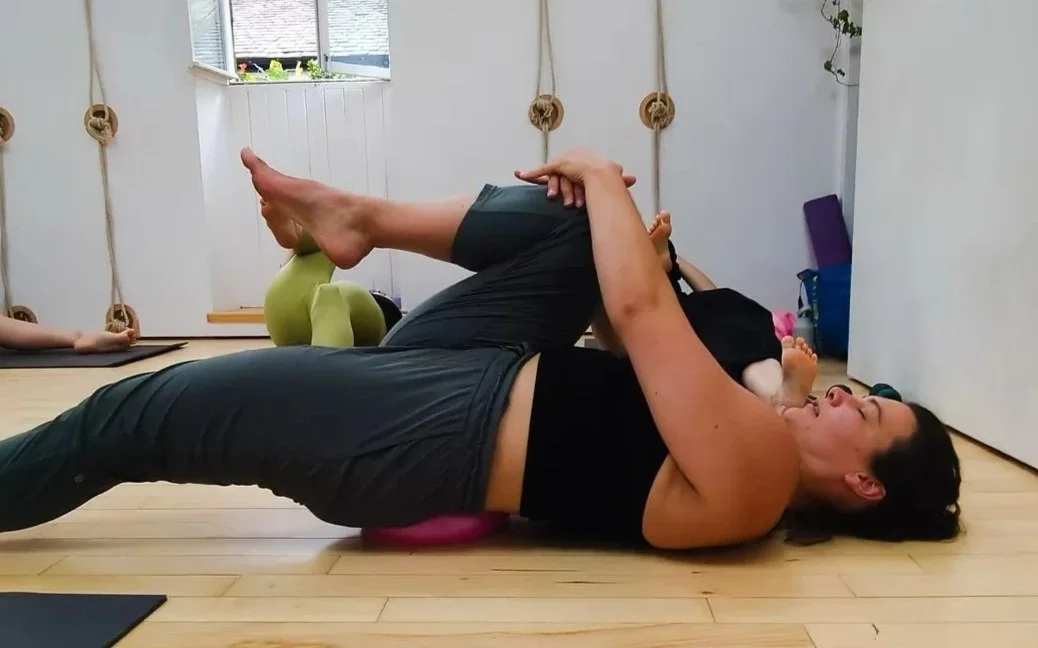 A woman practicing yoga indoors, lying on a mat with her eyes closed, holding her upper leg with both hands. She is wearing a black top and pants, with other yoga equipment like kettlebells and mats in the background.