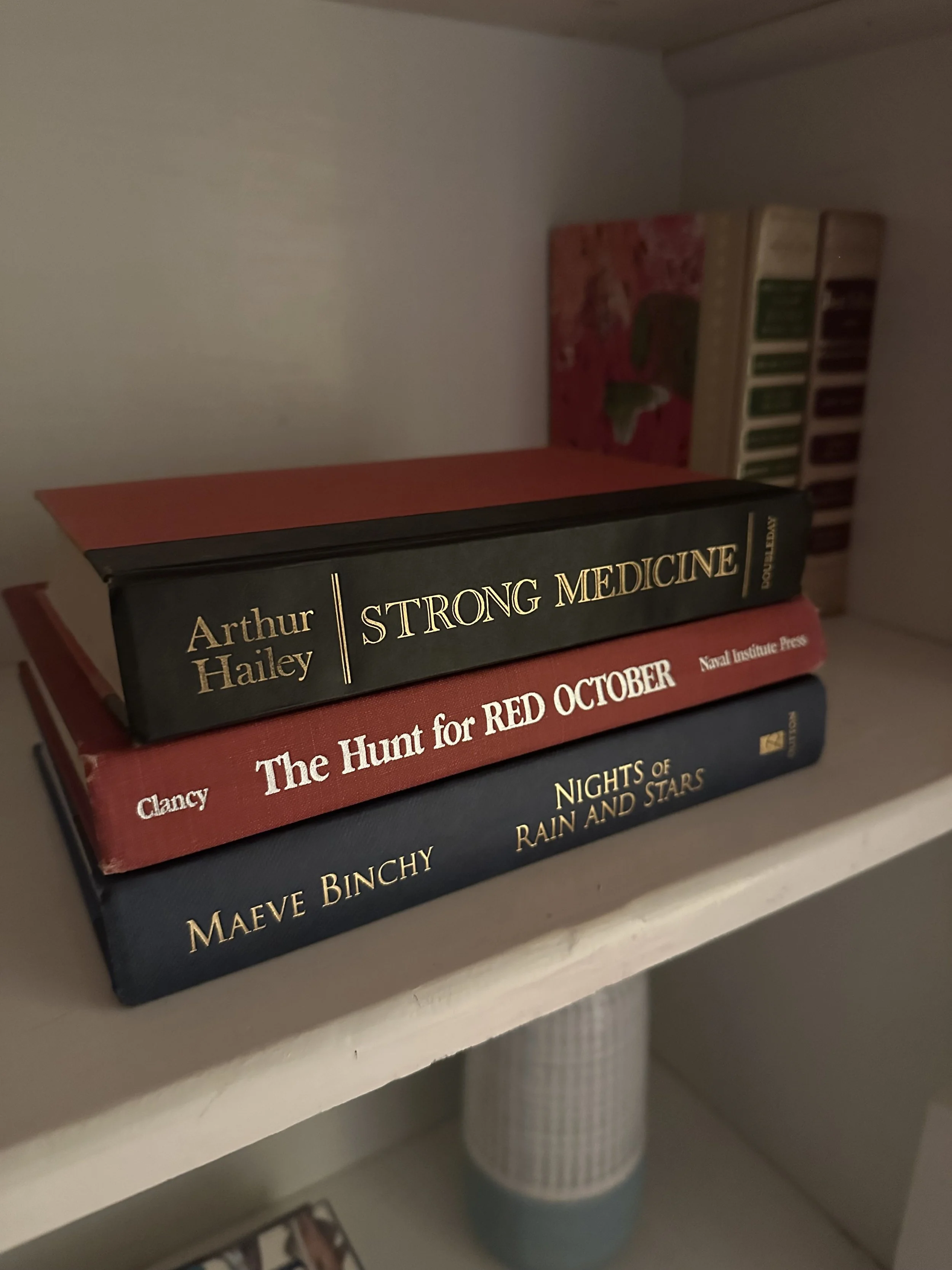 A stack of three books on a white shelf. Titles: "Strong Medicine," "The Hunt for Red October," and "Nights of Rain and Stars." Warm, cozy ambiance.