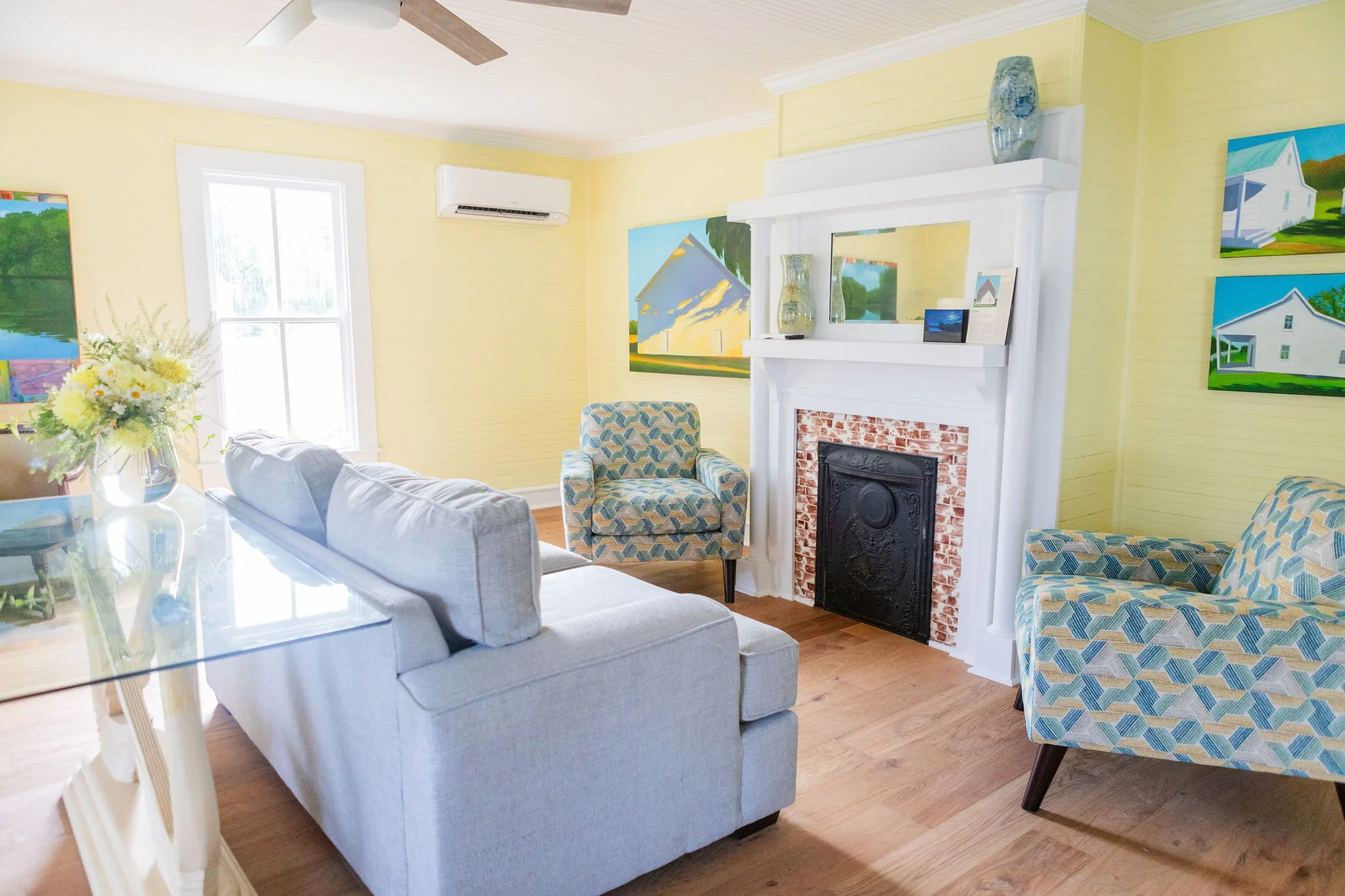 Bright living room with light yellow walls and wood floor. Features a gray sofa, two patterned armchairs, a decorative fireplace, and colorful wall art.