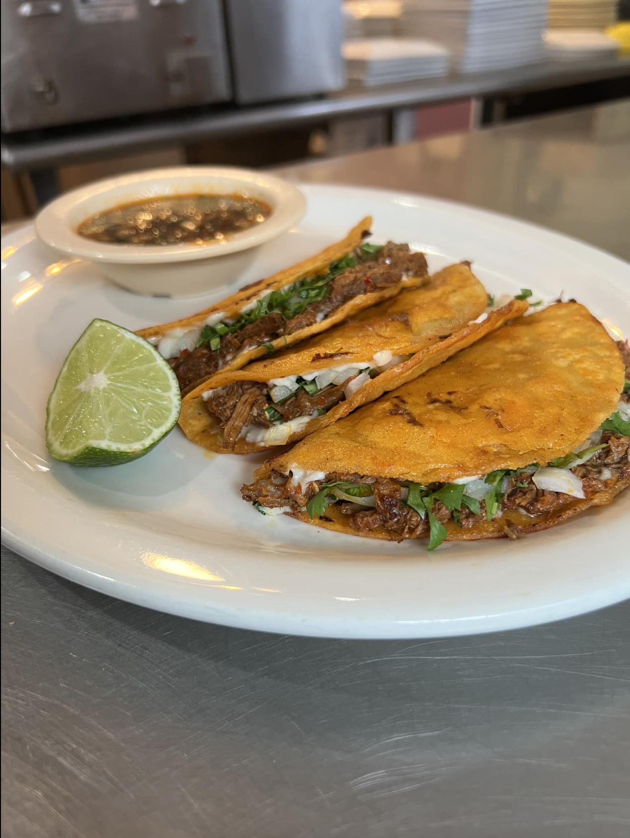 Three crispy birria tacos filled with beef, cilantro, and onions are on a white plate with a lime wedge. A bowl of dipping sauce is in the background.
