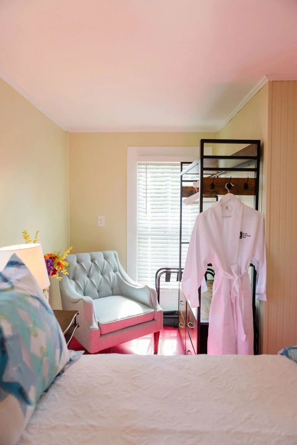 Cozy bedroom with a light blue armchair near a window. A white robe hangs on a black metal rack. Bed with colorful pillows in the foreground.