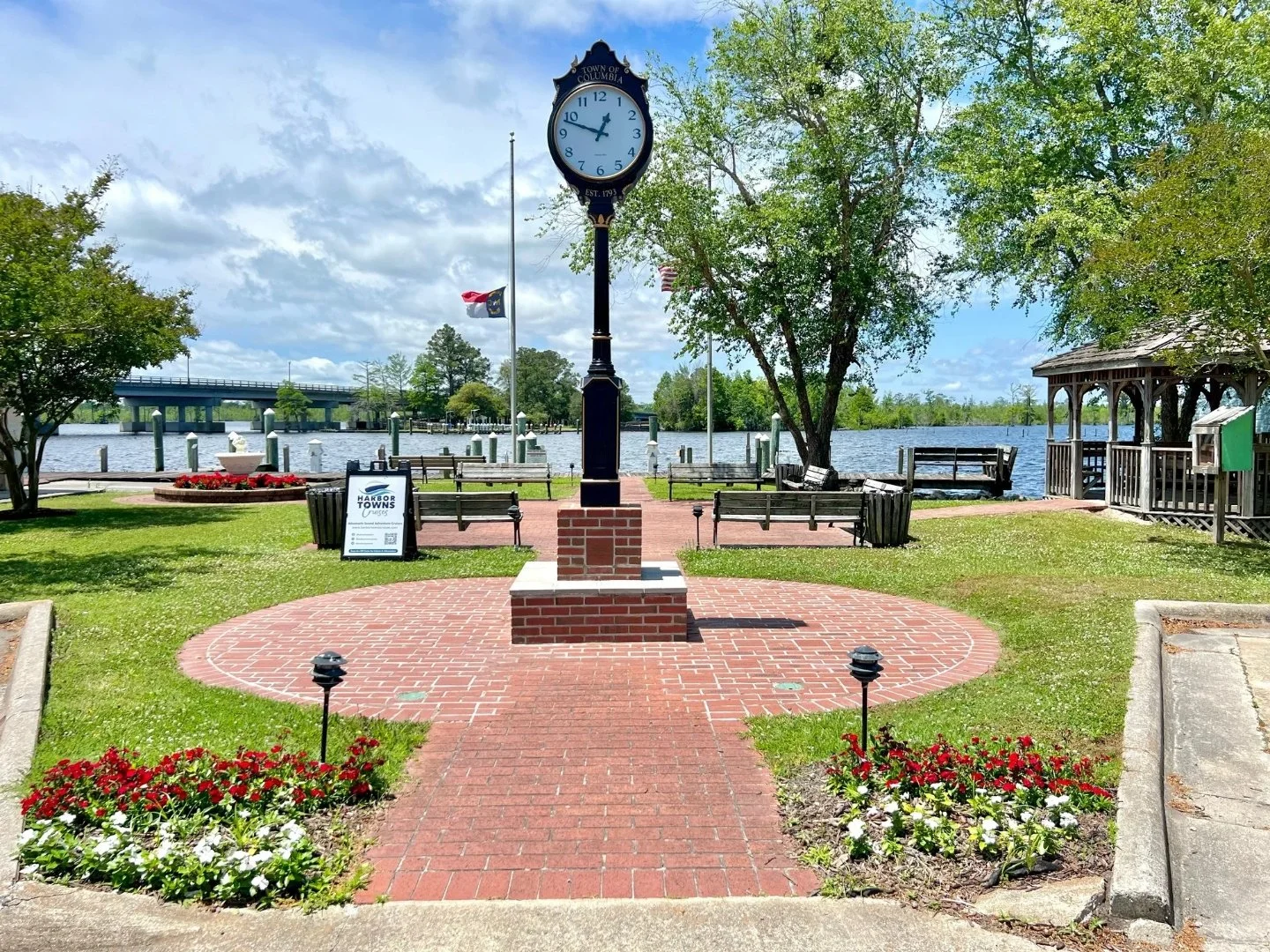 Columbia town square with red & white blooming flowers, clock in the center of the courtyard, view of the Scuppernong Riverwalk, and bridge