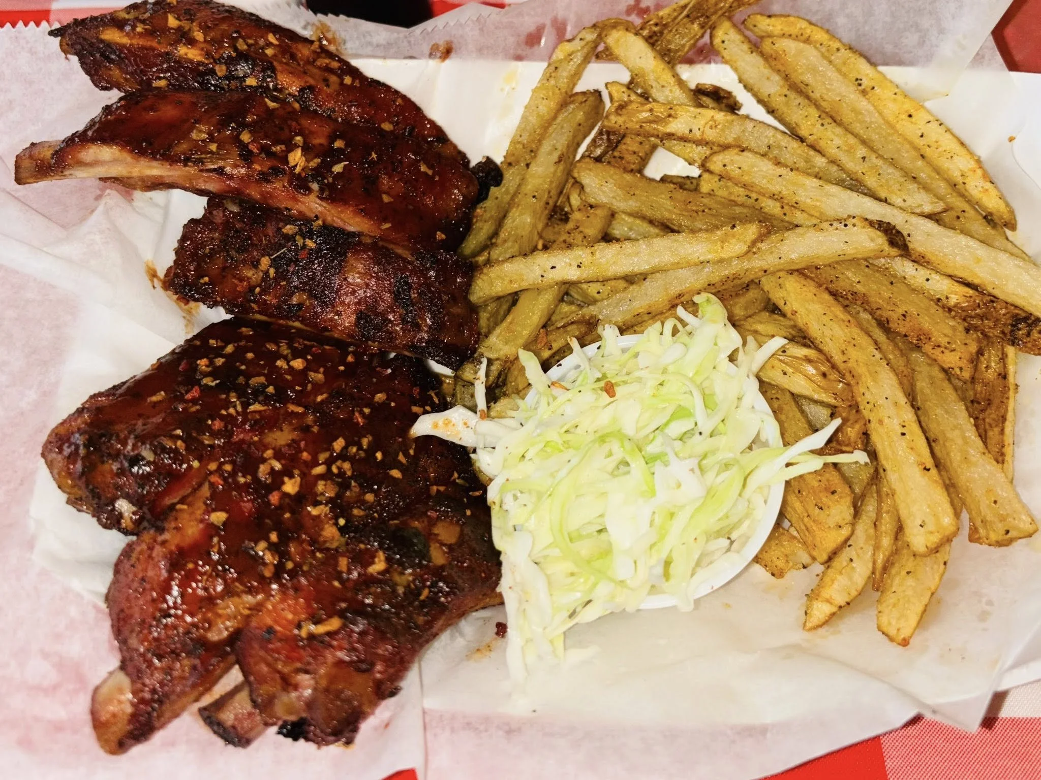 Grilled pork ribs garnished with spices, served with seasoned fries and a side of creamy coleslaw on a red checkered tablecloth, evoking a hearty meal.