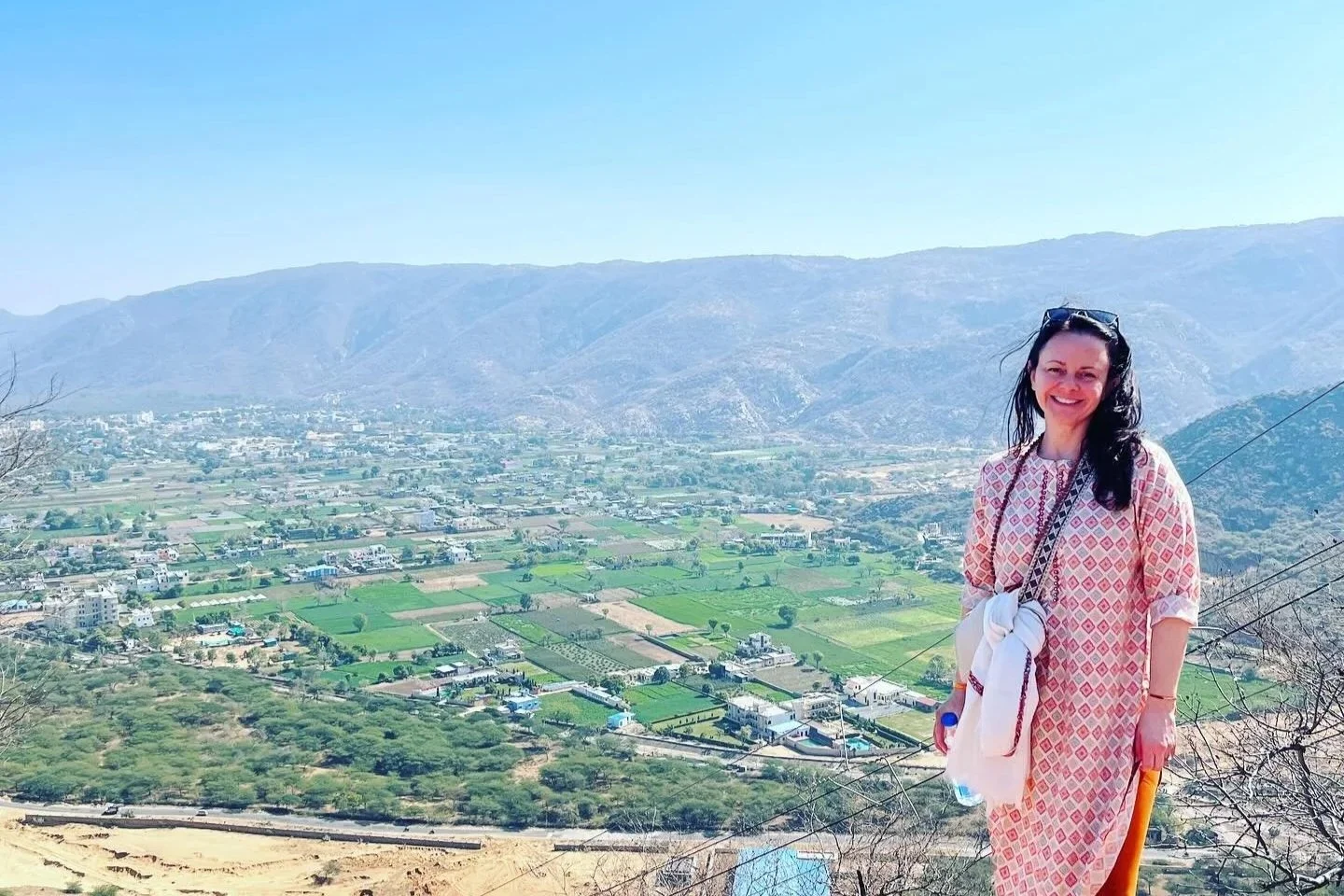 Heather Redabaugh in a patterned dress standing on a hill in Pushkar, India with a scenic view of a valley, fields, houses, and mountains in the background.