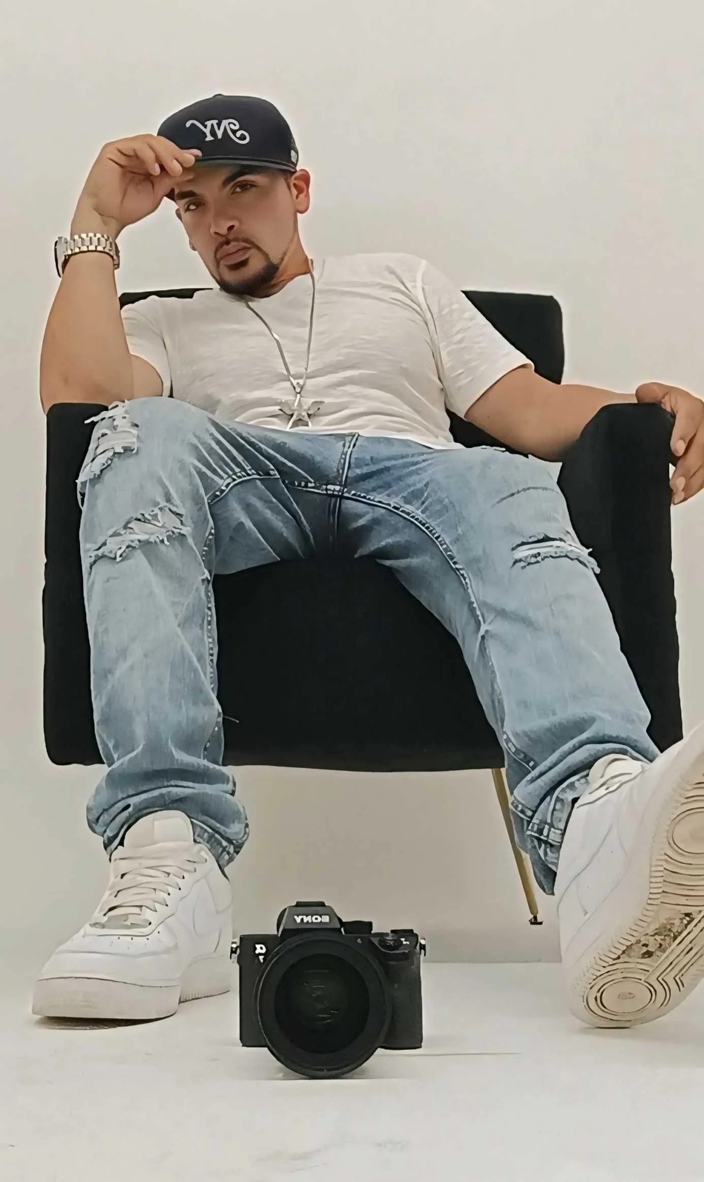 Person wearing a cap, white shirt, ripped jeans, and white sneakers sitting in a chair, with a Sony camera on the floor in the foreground.