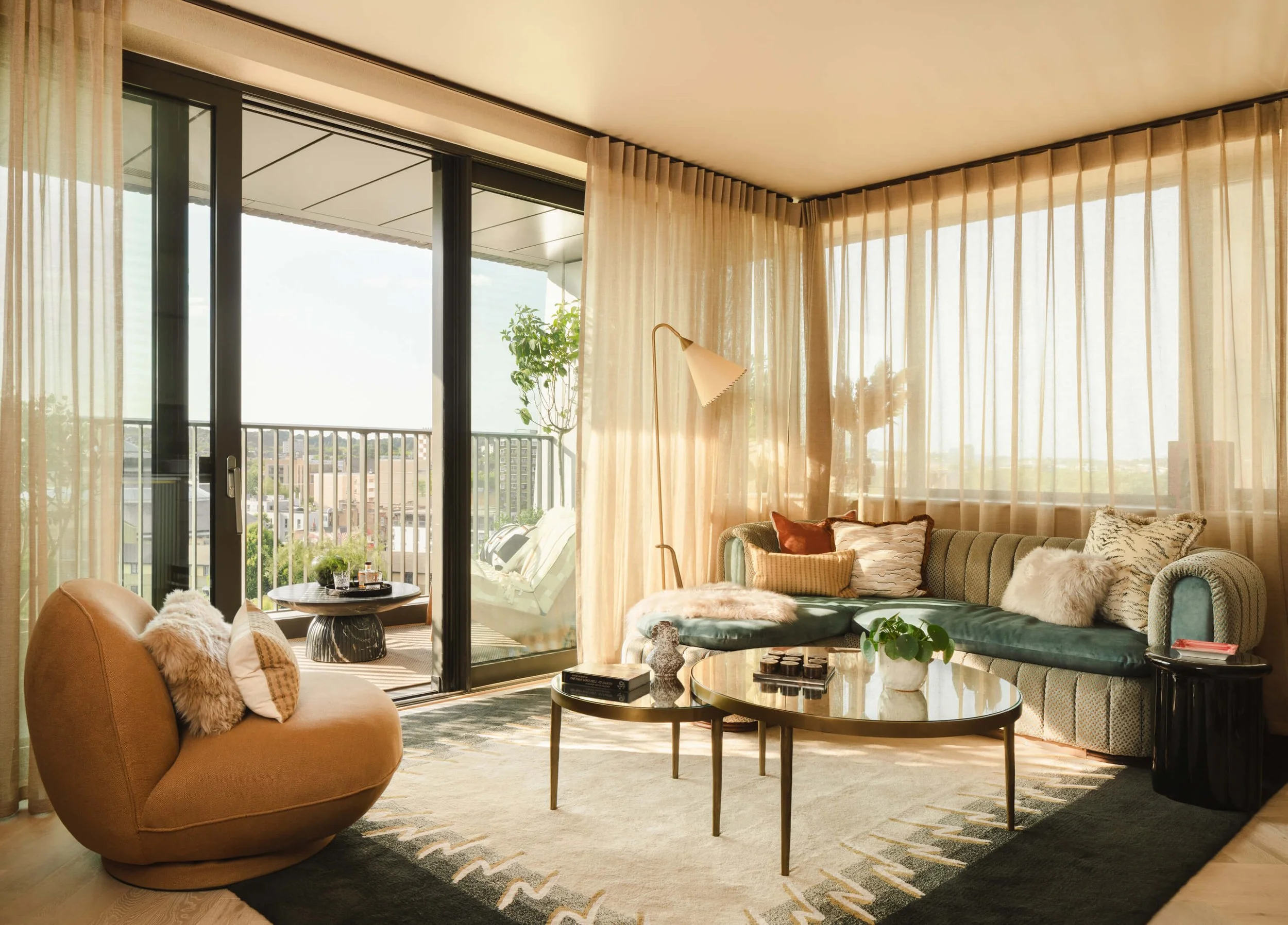 Living room with glass sliding door leading to balcony, beige curtains, velvet sofa with throw pillows, round coffee table with books and plant, cozy chair with pillows, potted tree, and floor lamp, during daytime.