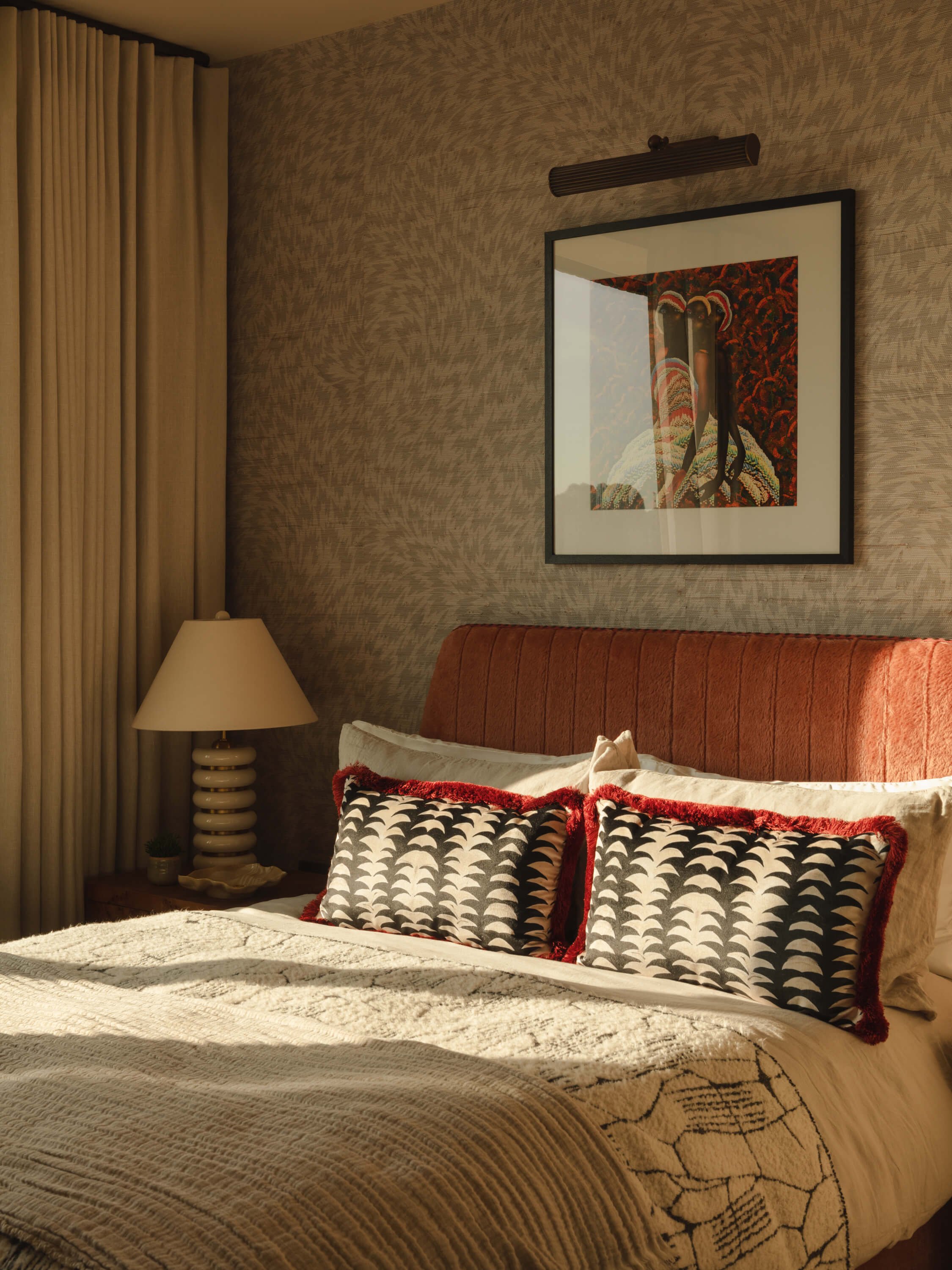A bedroom with a bed, black and white patterned pillows with red trim, a beige textured bedspread, a bedside lamp, a framed colorful artwork of an African tribal mask and figure on the wall, and cream-colored curtains.