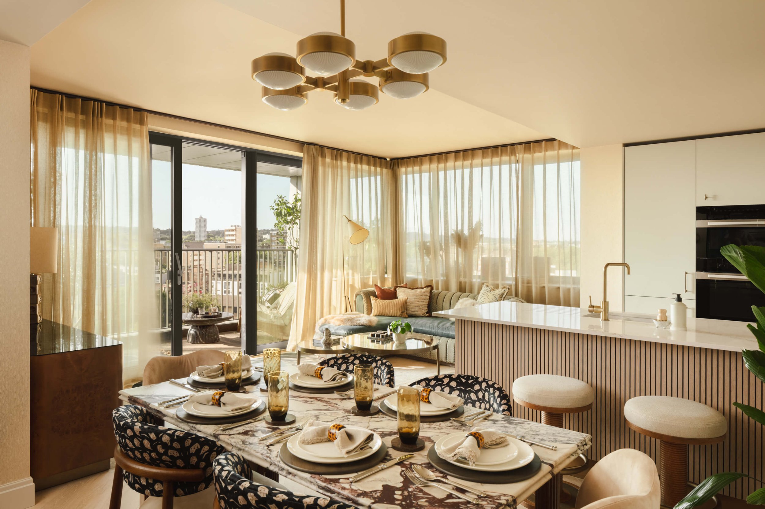 Modern living and dining area with a marble dining table set for six, bohemian patterned chairs, a cozy sofa near large windows, and a white kitchen island with a sink, gold faucet, and bar stools, illuminated by soft natural light.