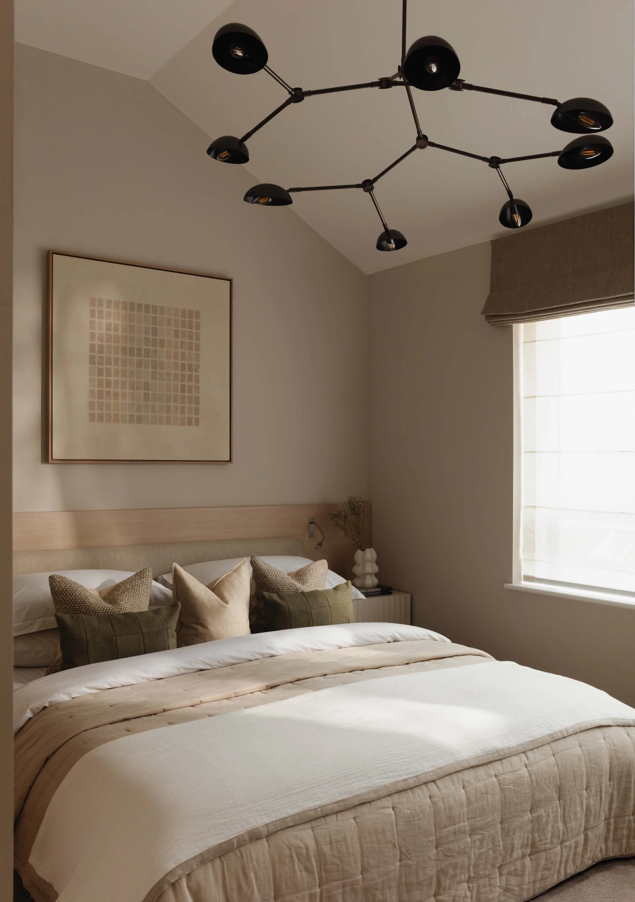A neatly made bed with beige bedding and multiple pillows in a bedroom with a modern black ceiling light fixture, a framed abstract art piece, and a window with a beige Roman shade.