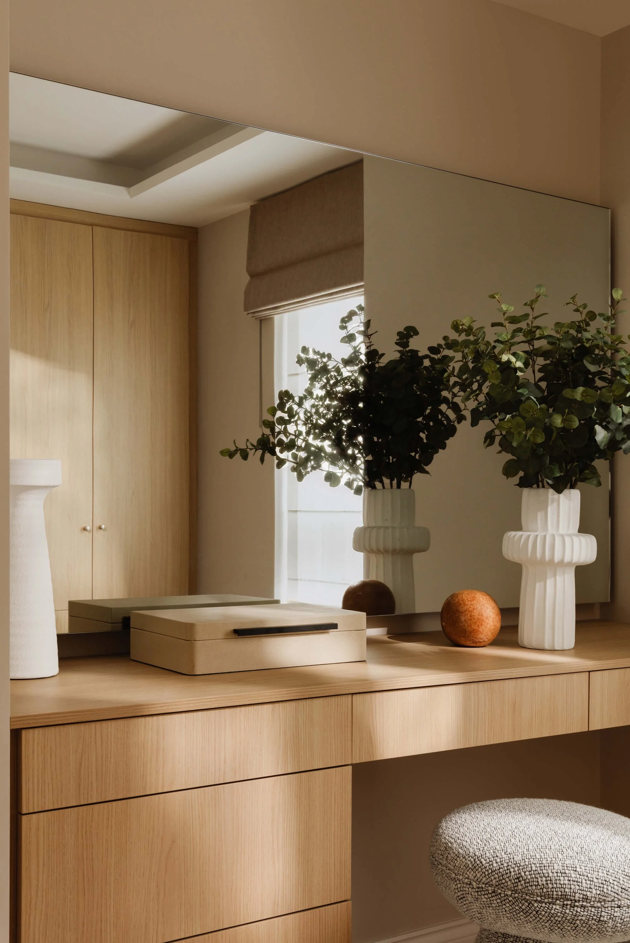 A wooden vanity table with a large mirror, decorated with two white vases containing green foliage and two round decorative objects, with a window in the background.