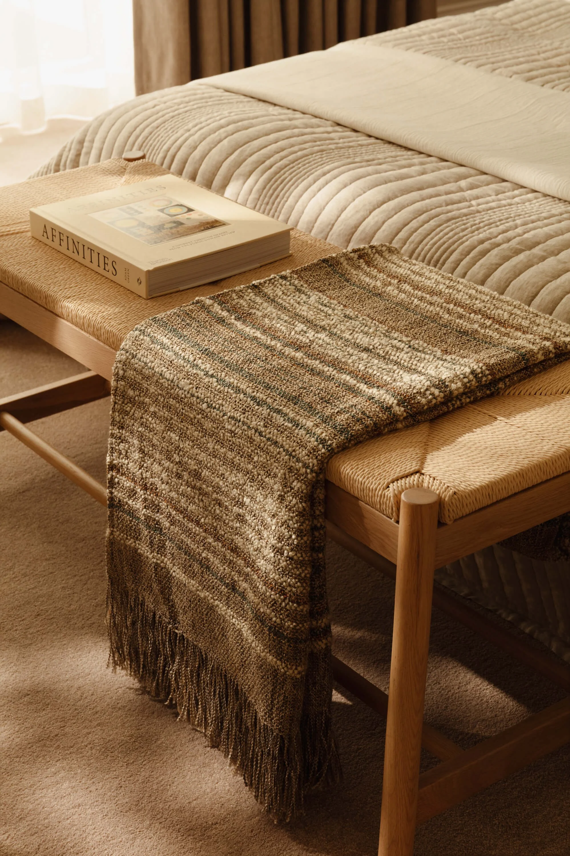 A cozy bedroom scene featuring a wooden bed with a beige quilted blanket, a multicolored woven throw draped over the side, and a tray table with a book titled "Affinities" on top.