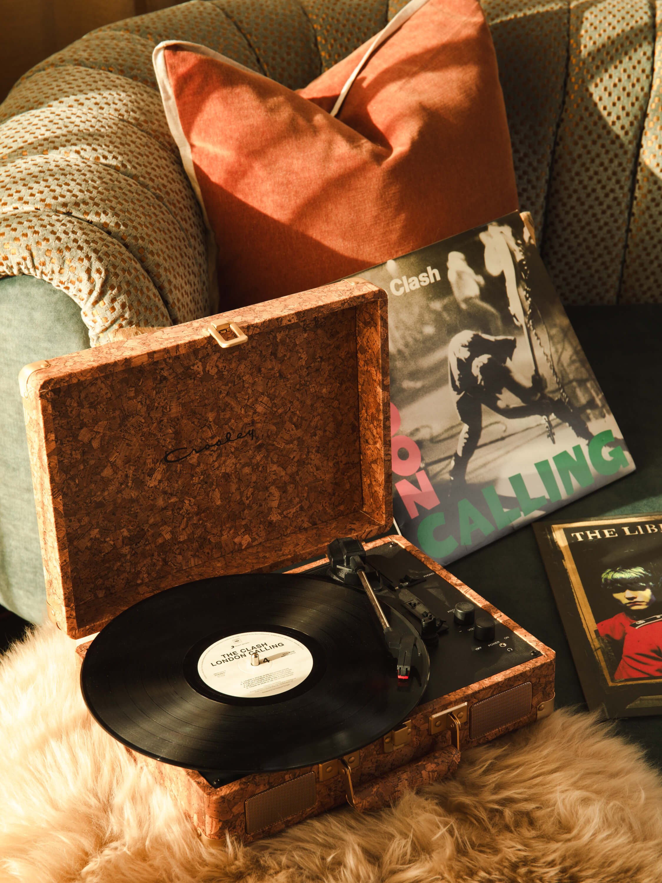 A vintage portable record player with a vinyl record spinning on it, surrounded by vinyl albums including 'London Calling' by The Clash and 'The Libertines.' A plush pillow and a textured sofa are in the background, with a fur rug beneath the record 
