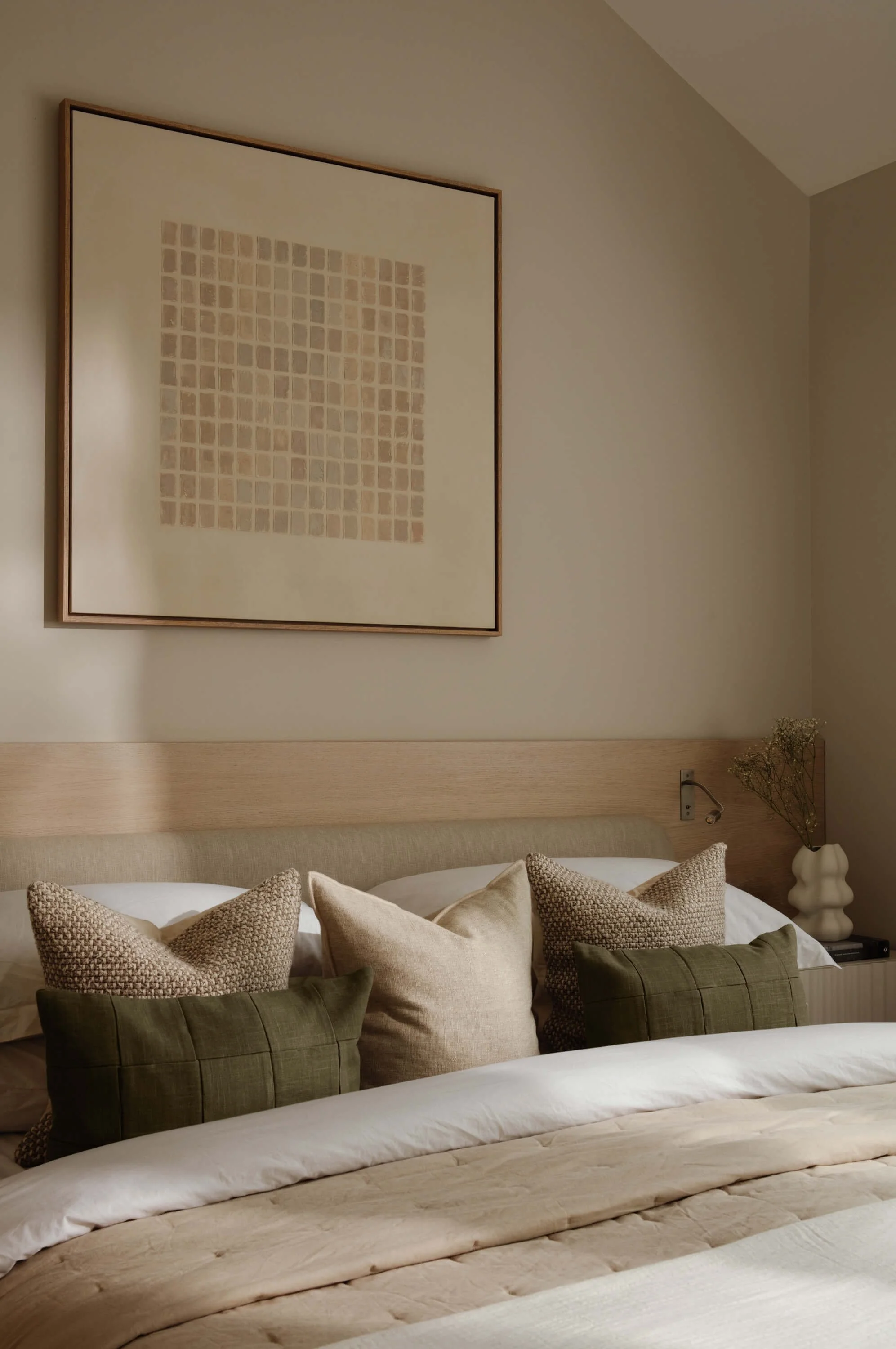 A neatly made bed with multiple throw pillows in beige and green, positioned against a light-colored wall with a framed abstract artwork. A bedside table with a vase and dried flowers is visible on the right.