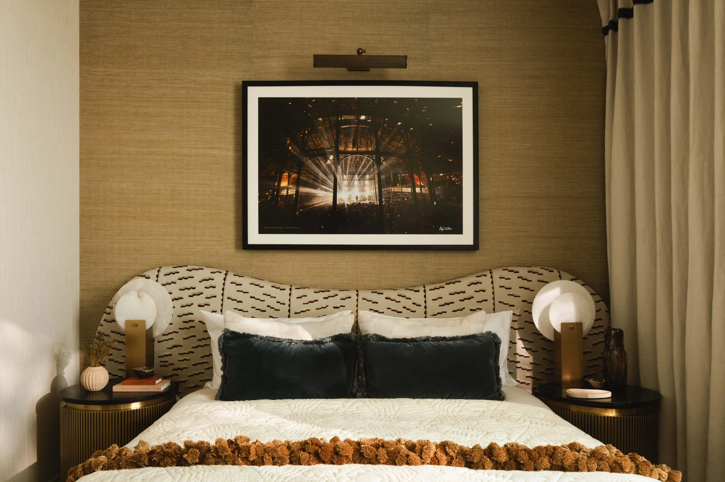 A hotel bedroom with a patterned headboard, two nightstands with lamps, books, and decorative items, pillows on the bed, a framed concert photo on the wall, with curtains on the right side.