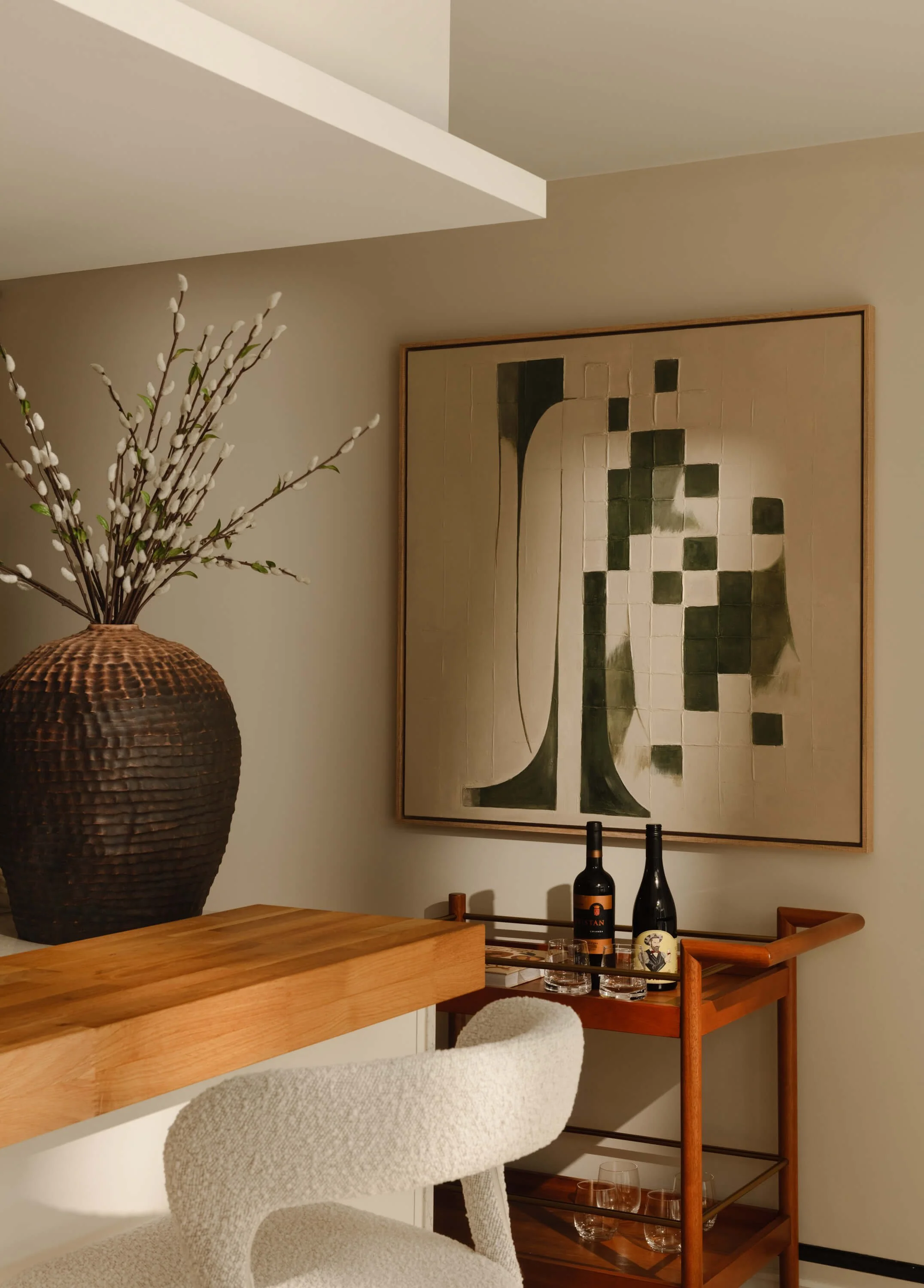 A corner of a living space with a large abstract painting on the wall, a wooden bar cart with bottles and glasses, a large textured vase with decorative branches, and a cushioned chair.