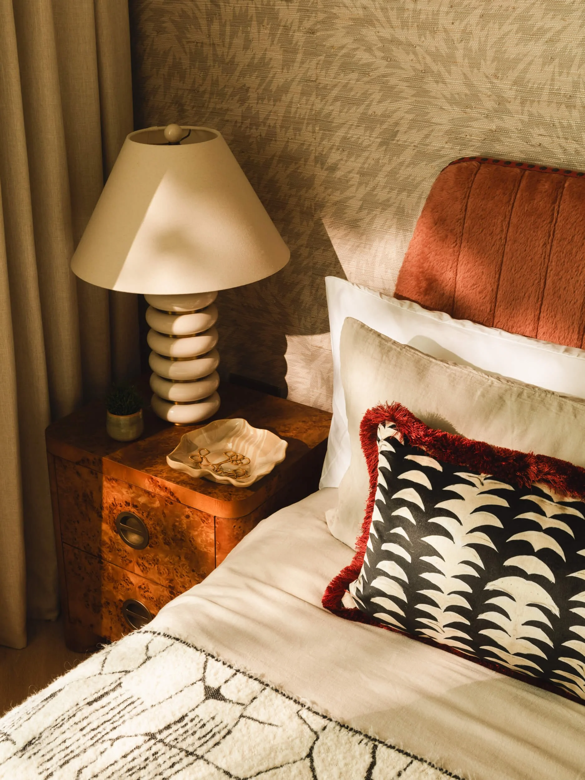 Bedroom corner with a wooden nightstand, a white ceramic dish with gold jewelry, a beige lampshade with a stacked sphere base, a small potted plant, a bed with white linens, a large white pillow, a decorative cushion with a black and white pattern fr