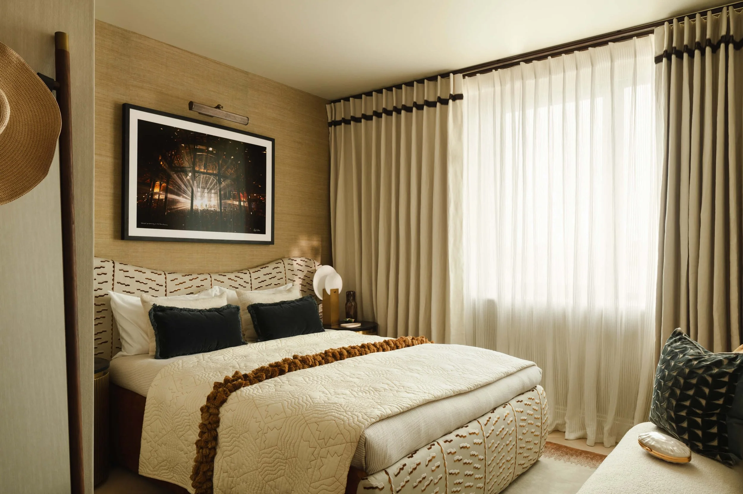 A hotel bedroom with a bed featuring a cream quilt and black pillows, a window with cream curtains, a nightstand with a white lamp, and wall art depicting sunlight streaming through trees.