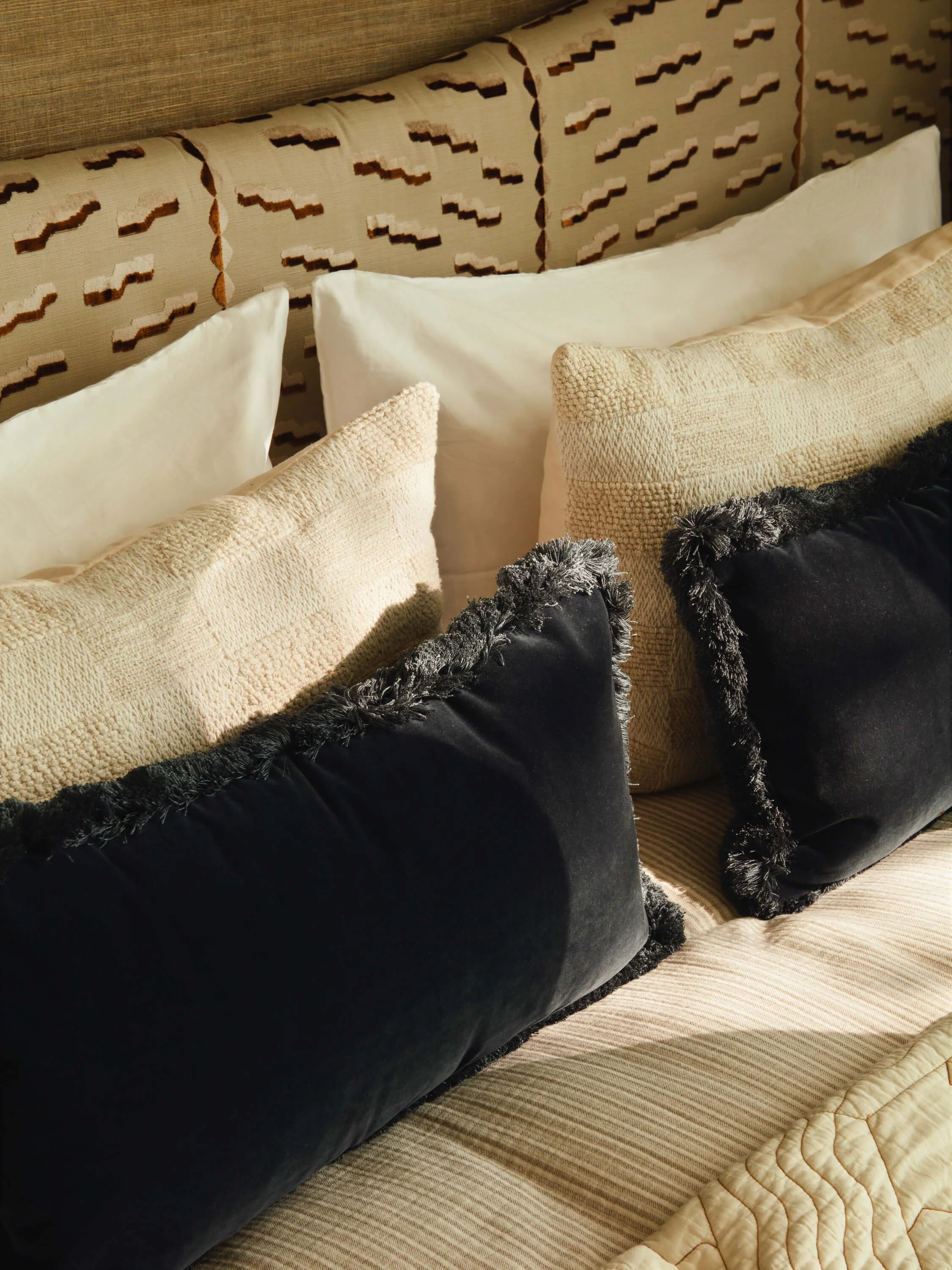 A collection of pillows in neutral colors on a bed, with a patterned headboard in the background and sunlight casting shadows.
