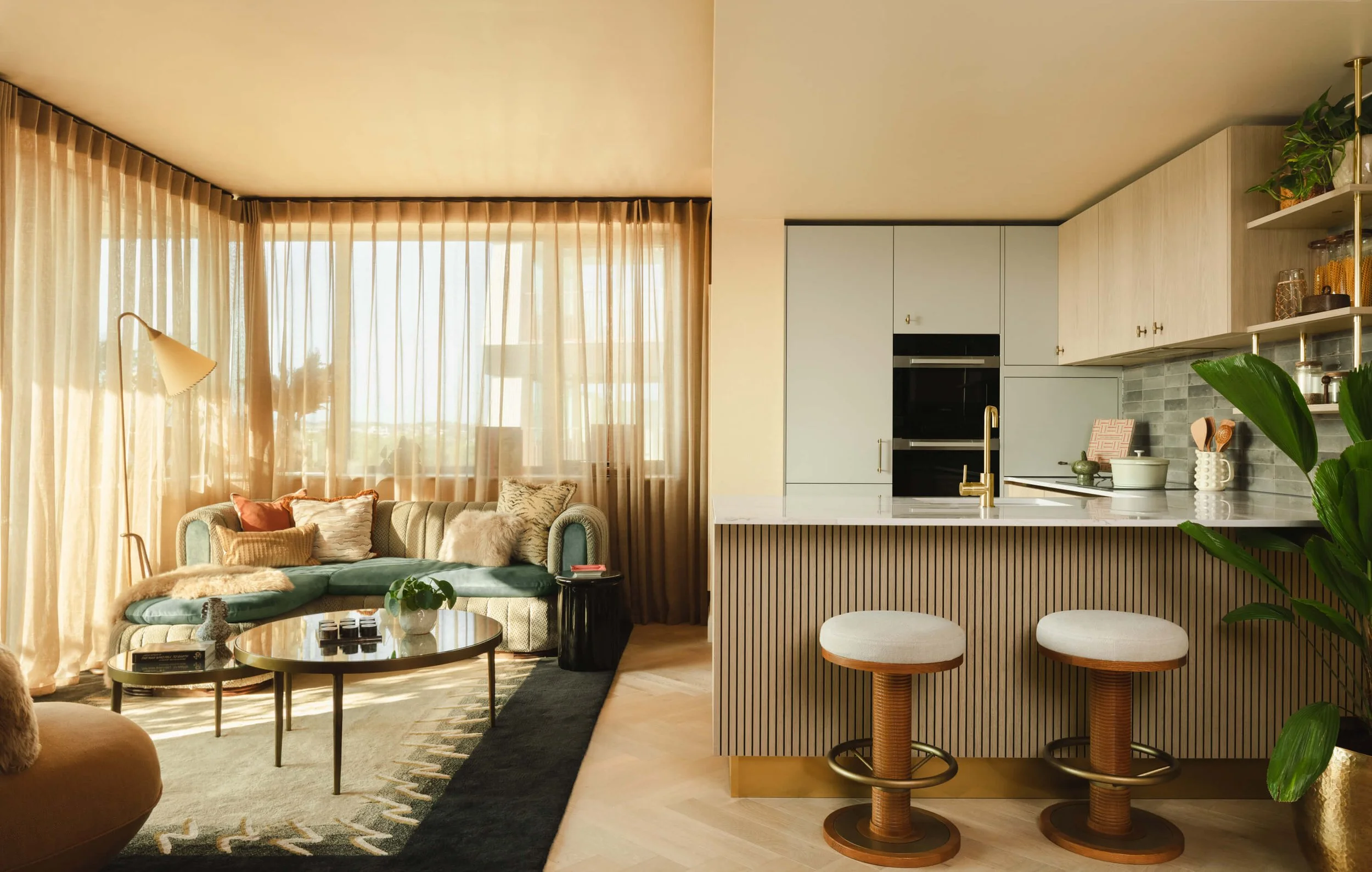 Living room and kitchen with beige curtains, green sofa with pillows, coffee table, bar stools, and modern kitchen cabinets with decorative items.