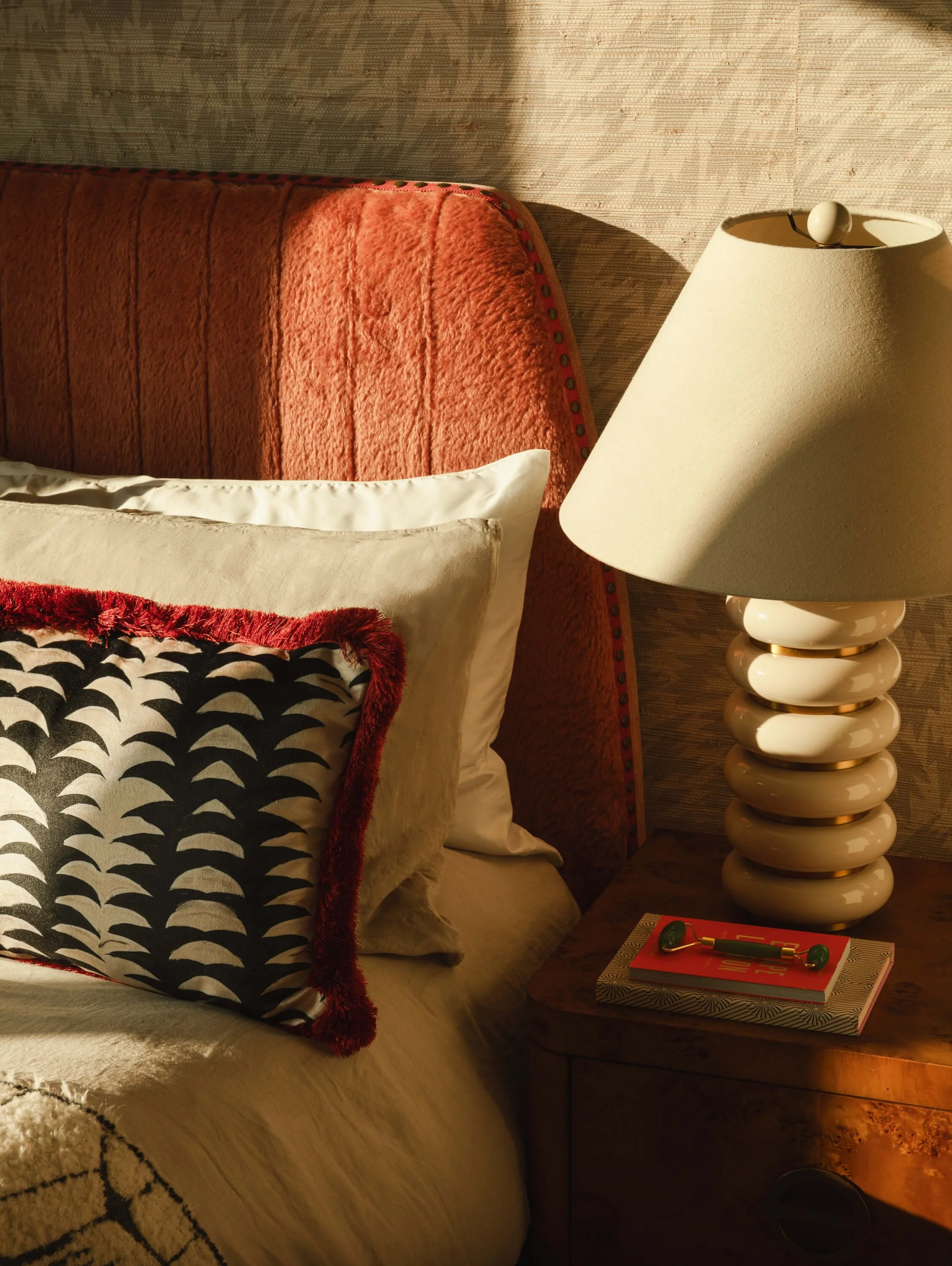 A bedroom corner with a textured orange headboard, cream and patterned pillows on a beige bedspread, a wooden nightstand with a modern striped lamp, a red book, and a small green object.