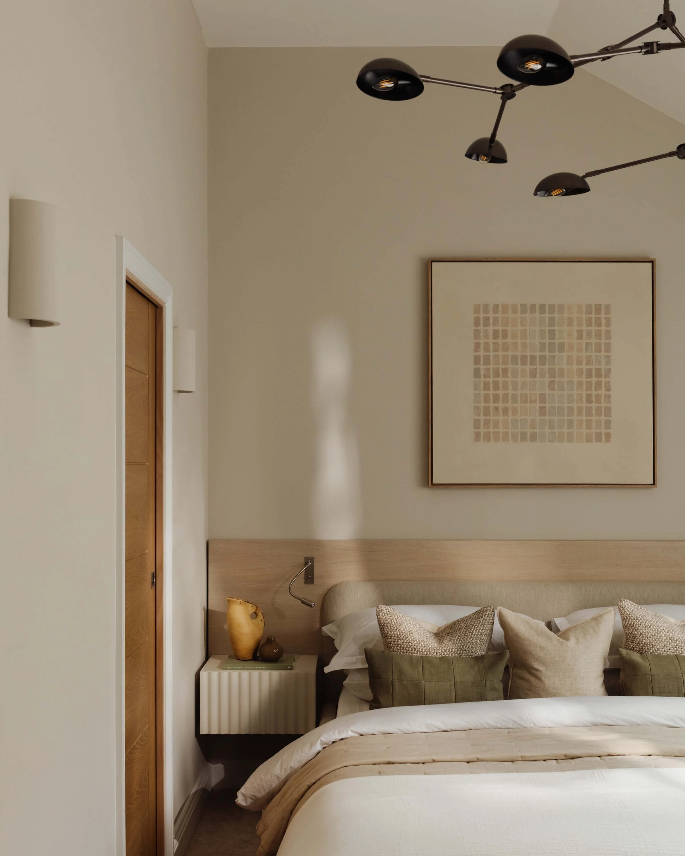 A bedroom corner with a bed, decorative pillows, a bedside table with vases, and a framed artwork on the wall.