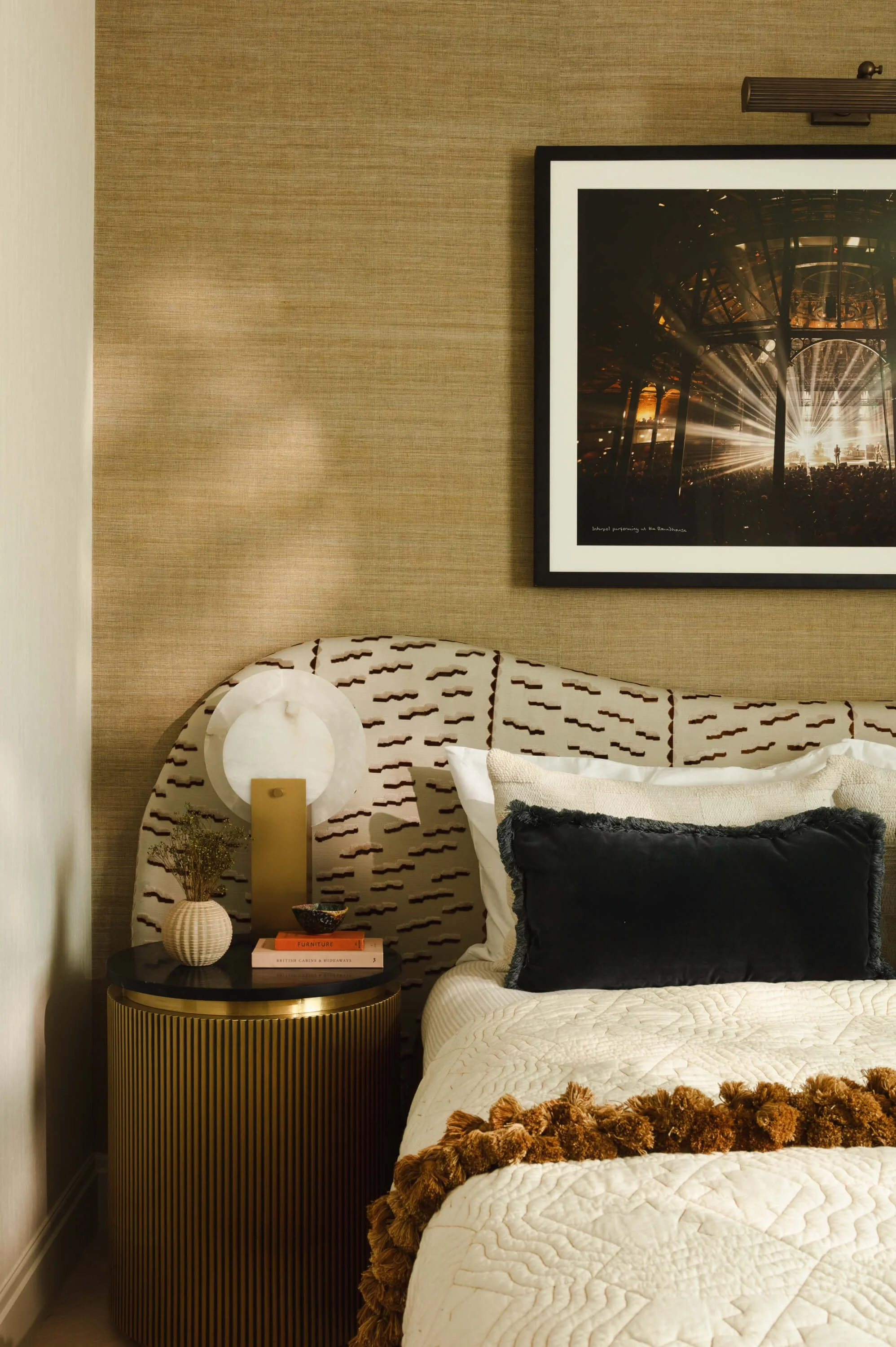 A bedroom corner featuring a bed with a textured cream quilt and a large black velvet pillow. Beside the bed is a round, gold-striped side table with decorative items, including a small vase with dried flowers, a white spherical lamp, and books. A fr