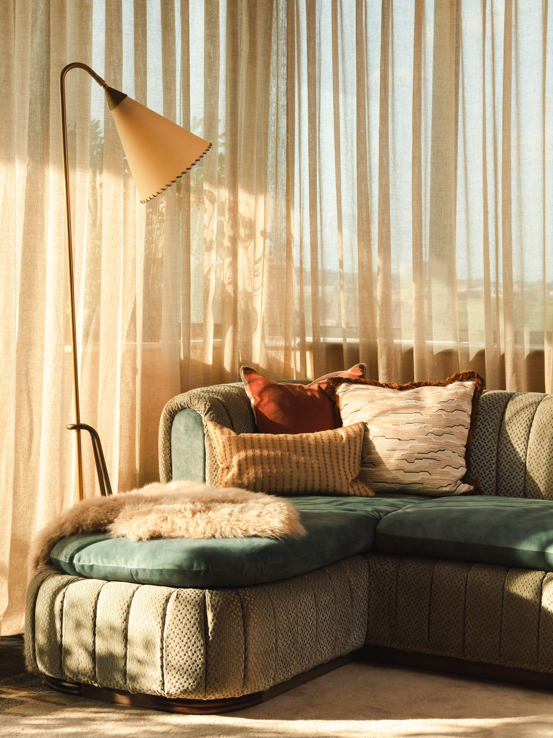 A living room with a green upholstered sectional sofa, decorative pillows, a sheepskin throw, and a tall floor lamp next to beige curtains letting in natural light.