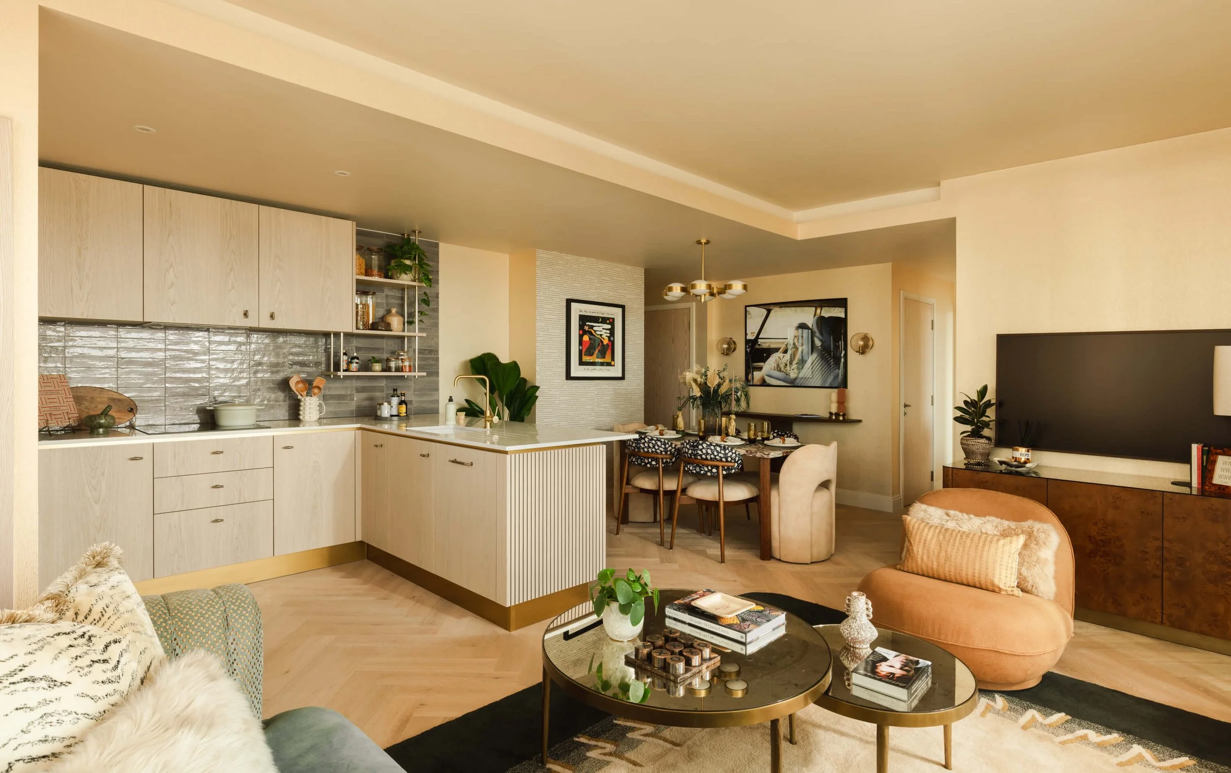 Open-concept living room and kitchen with beige and brown tones, featuring a curved beige couch with textured pillows, a modern black glass coffee table with books and decor, light wood flooring, a dining area with a round table and chairs, wall-moun