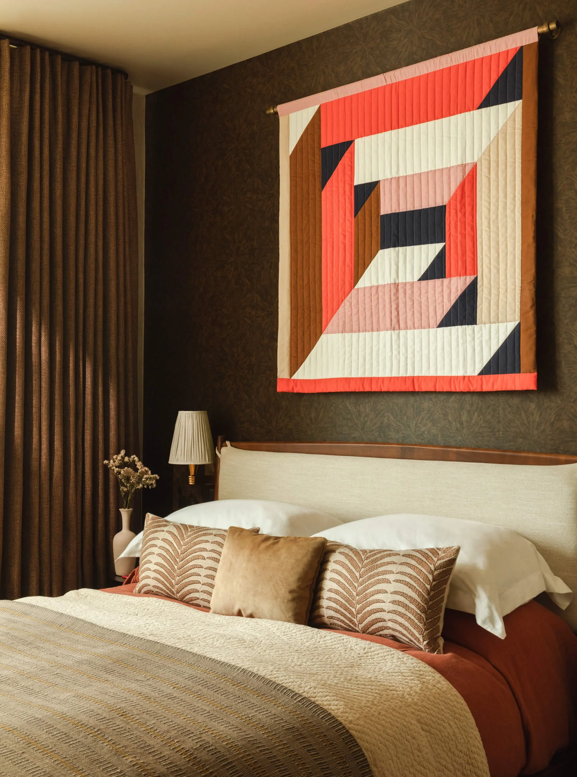 A bedroom with a bed featuring beige and rust-colored bedding and patterned pillows, a nightstand with a vase and flowers, a wall-mounted lamp, brown curtains, and a quilted abstract wall hanging in shades of red, coral, black, white, beige, and brow
