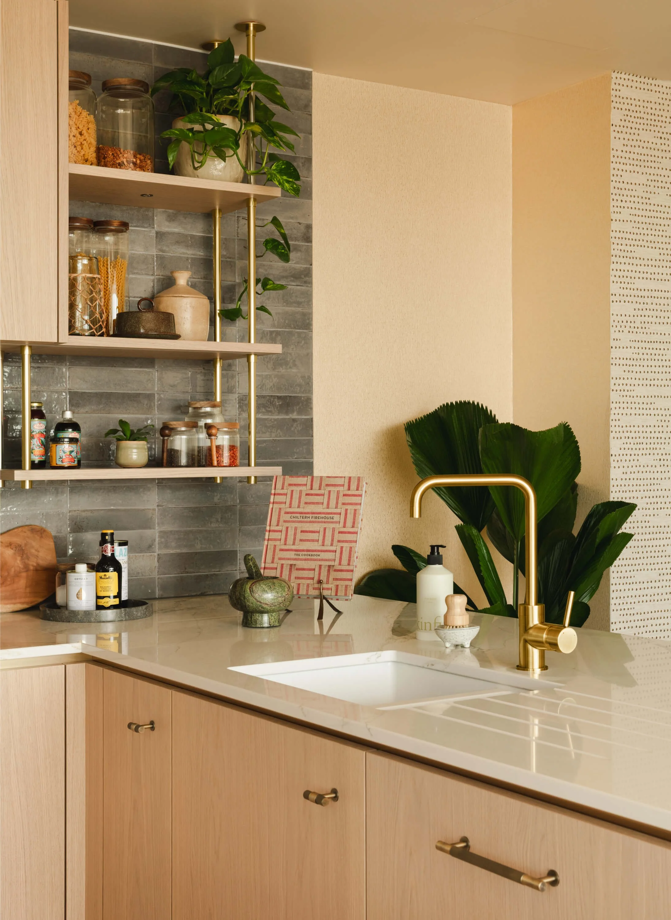 Modern kitchen with beige cabinets, white marble countertop, gold faucet, open shelving with jars and plants, green leaf decor, and various bottles and containers near the sink.