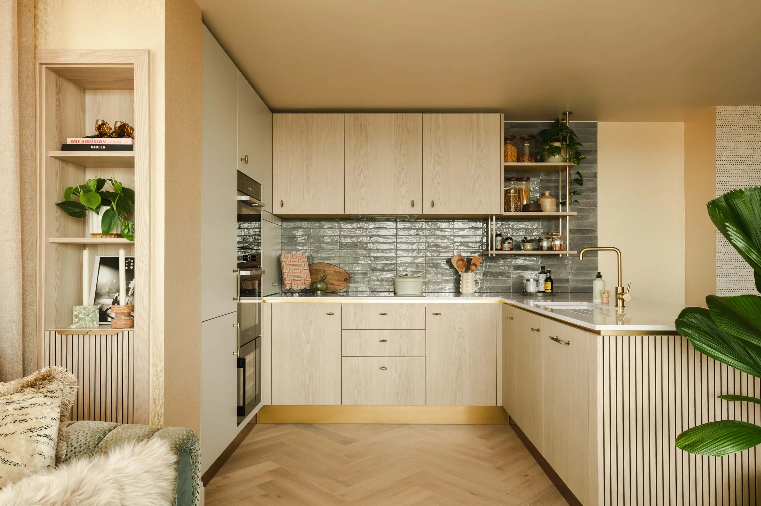 Modern kitchen with beige cabinets, black backsplash, and gold accents, featuring open shelves with jars and plants, a countertop with kitchen utensils, and a sink with a gold faucet.