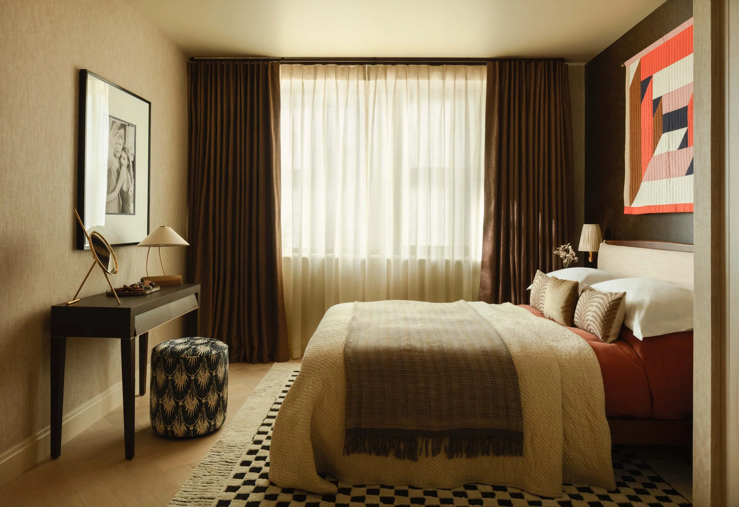A cozy bedroom with a large window, beige curtains, a bed with pillows, a nightstand with a lamp, a framed black and white photo on the wall, and a small table with a mirror and a patterned stool.