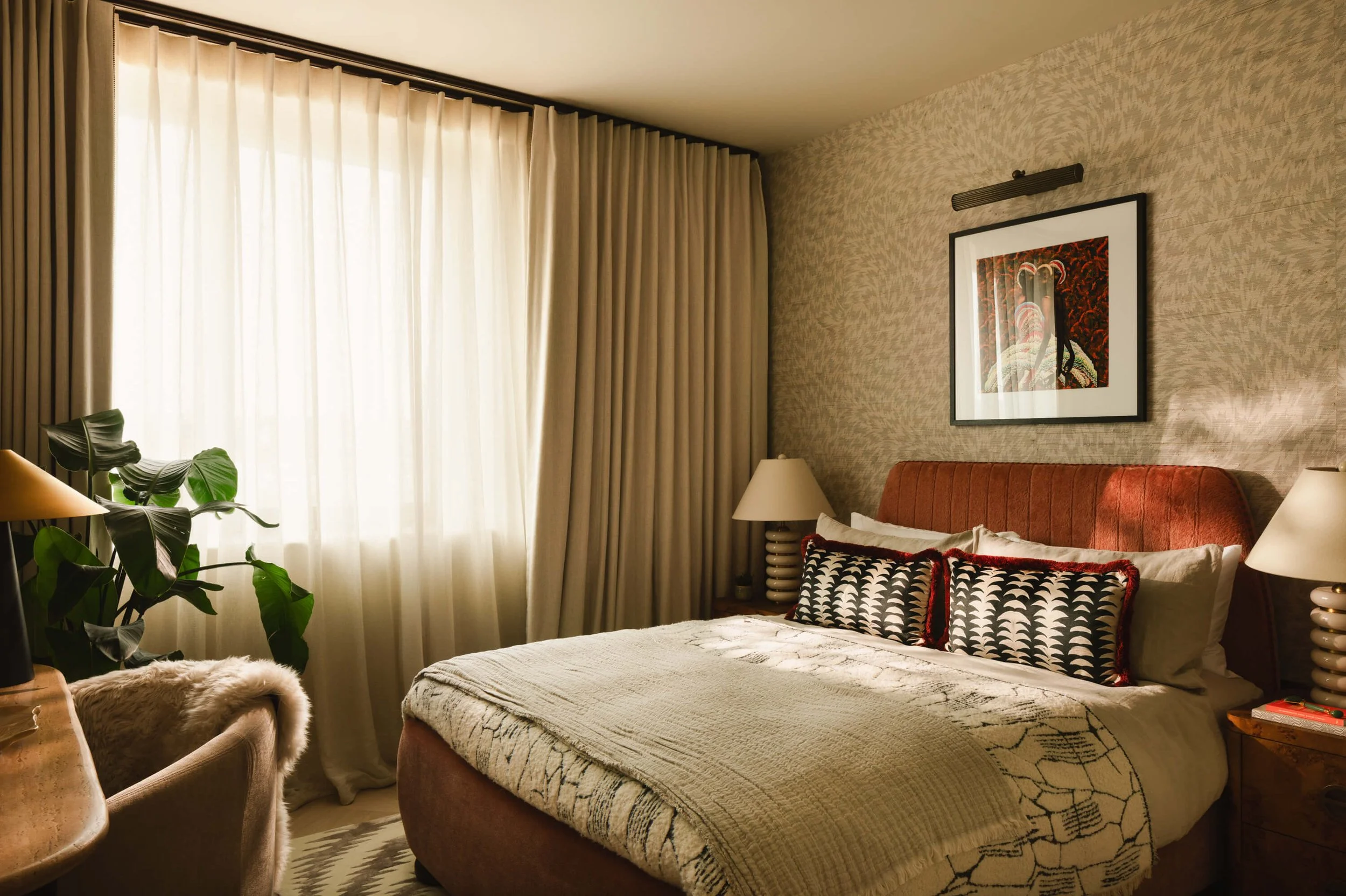 A cozy bedroom with a large bed, beige bedding, decorative pillows, and a burnt orange upholstered headboard. There are two nightstands with lamps on either side of the bed, a framed artwork above the headboard, a window with cream-colored curtains l