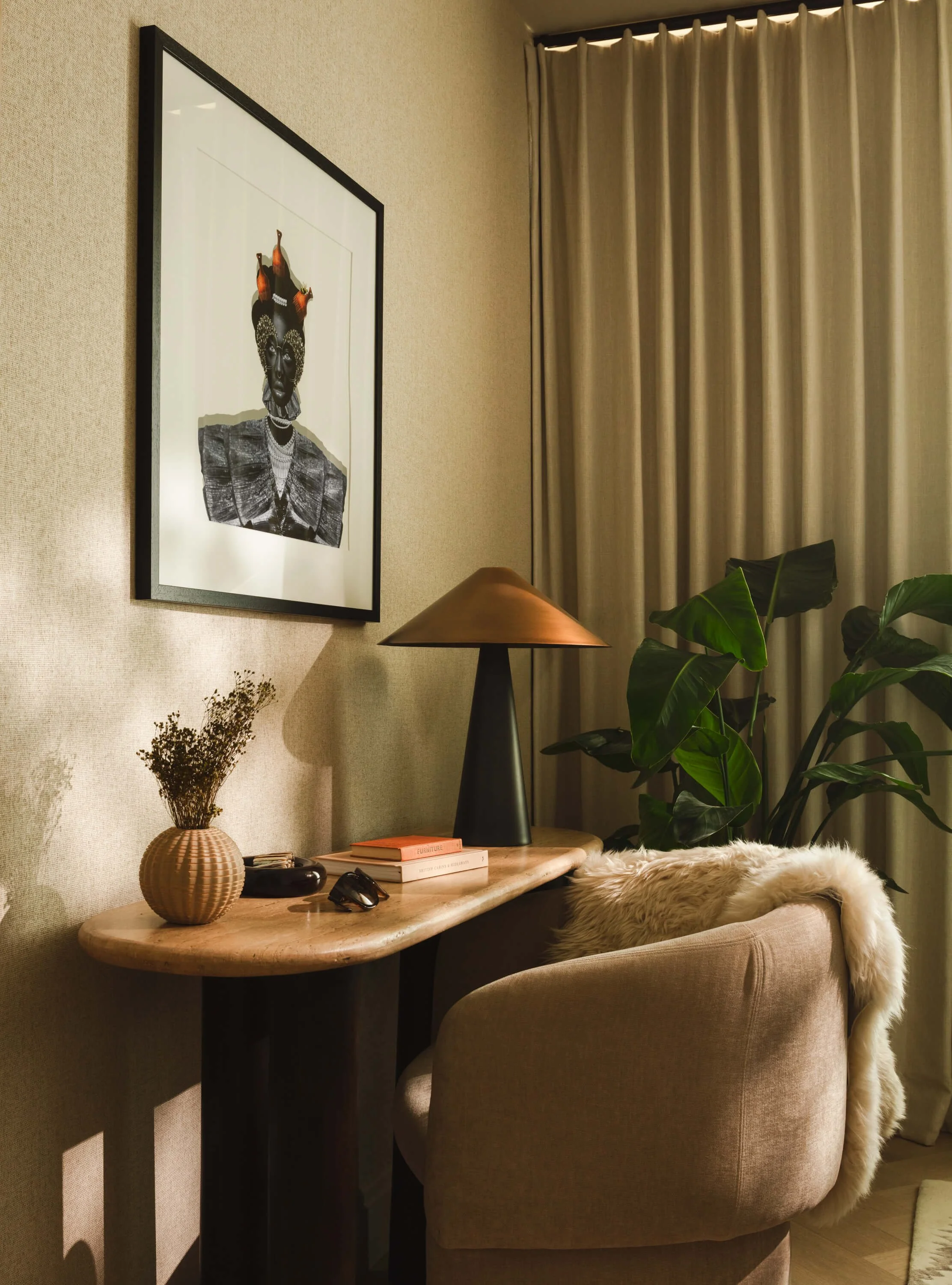 Interior of a cozy room with a wooden desk, beige chair with a fluffy throw, a table lamp, decorative vase with dried flowers, books, sunglasses, and a black-and-white framed artwork on the wall; a large green plant and curtains in the background.