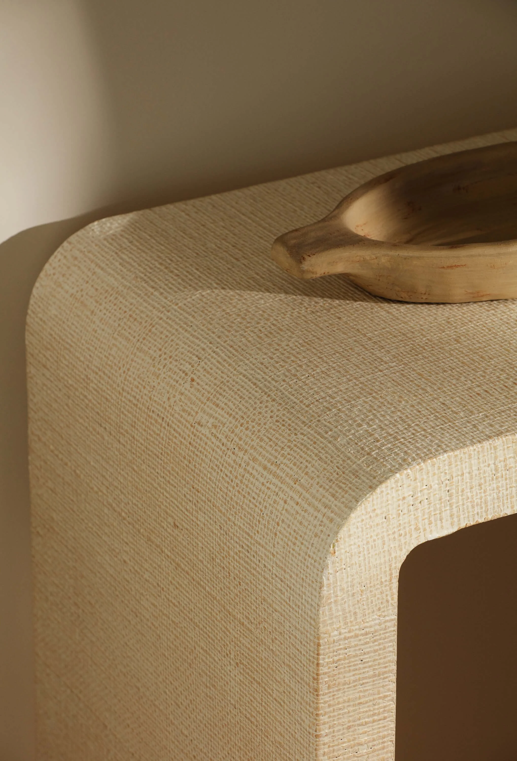 Close-up of a textured beige table with a curved edge, topped with a wooden bowl placed on the right side, against a plain wall background.