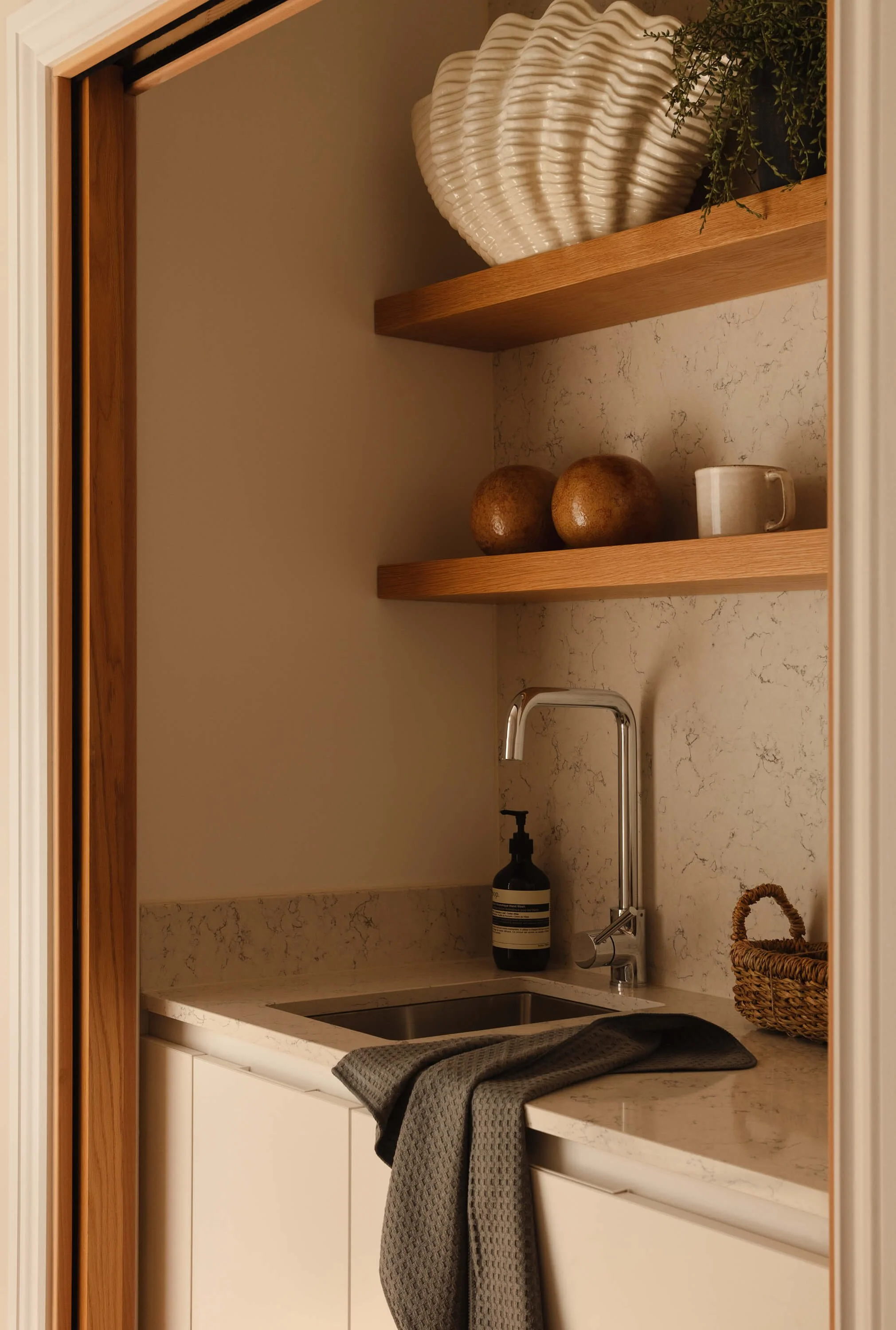 A small kitchen or utility space with a marble countertop, a stainless steel sink, a black soap dispenser, a white cabinet, and two wooden shelves. The shelves hold a large shell-shaped vase, two round brown objects, a white mug, and a small potted p