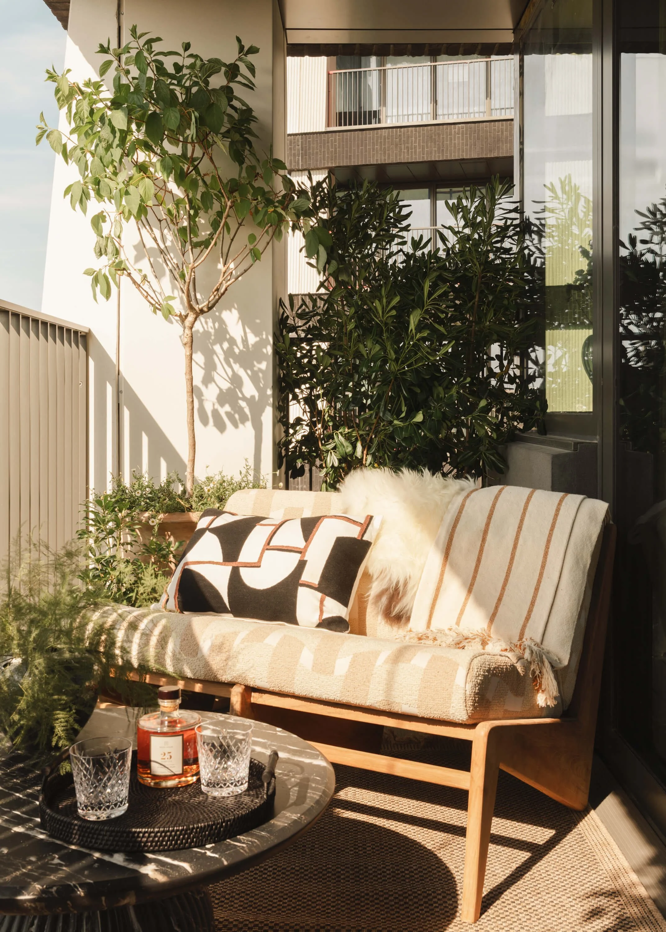 Sunlit balcony with a wooden sofa, patterned cushions, potted plants, and a small table with a bottle and glasses.