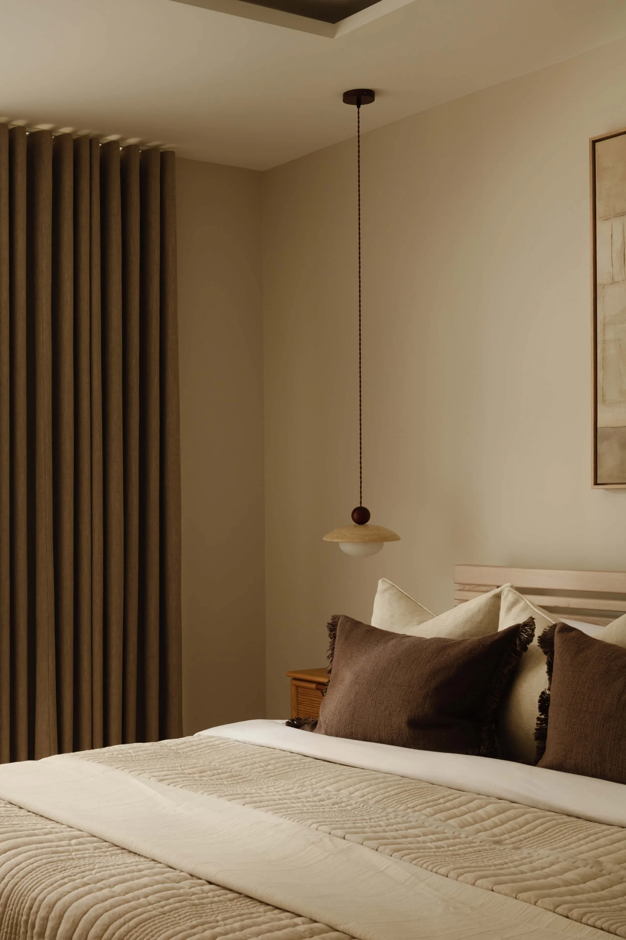 A neatly made bed with beige and brown pillows, a beige bedspread, and a small wooden nightstand next to it. A modern hanging pendant lamp is above the bed, and beige curtains are drawn closed on the window to the left.
