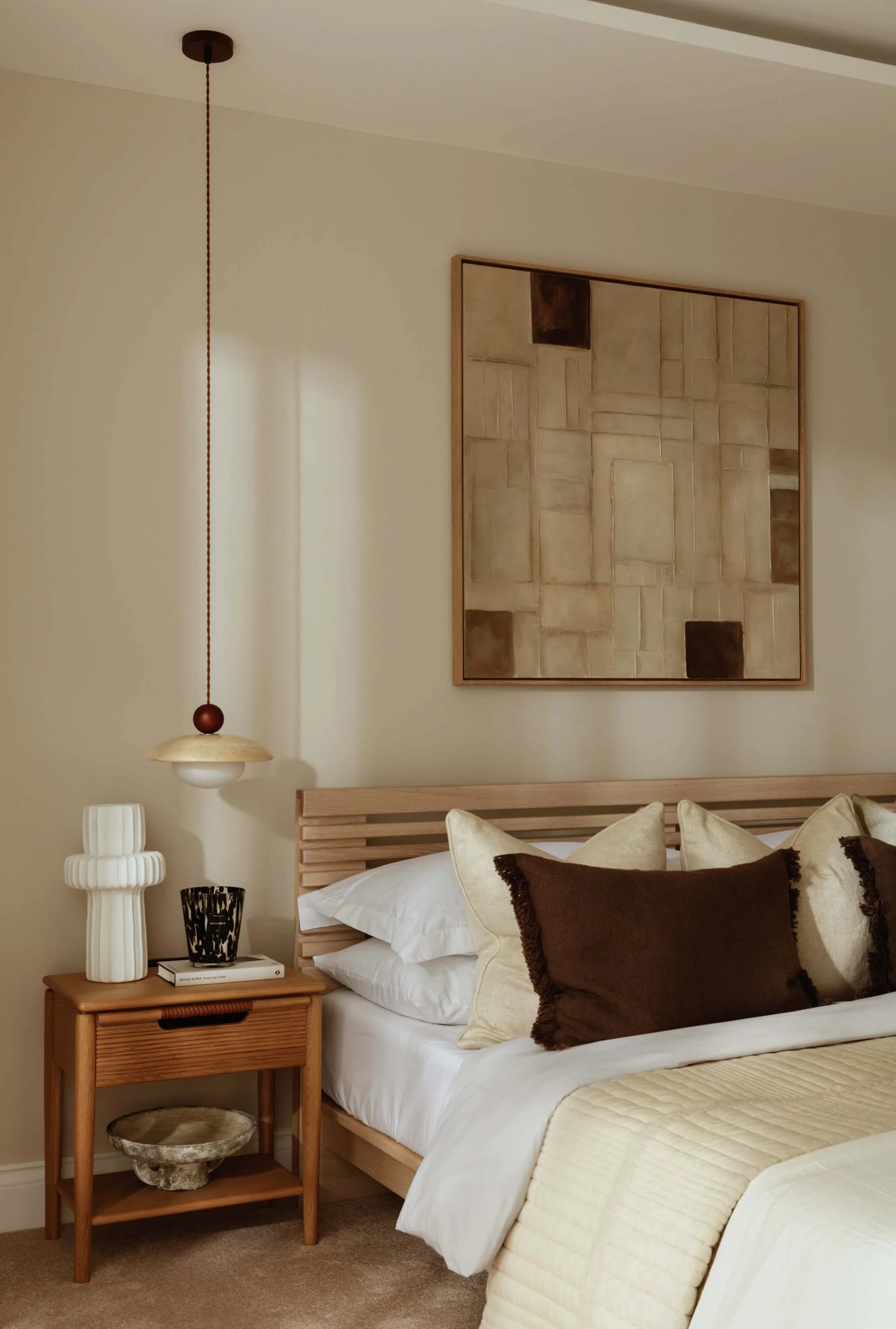 A bedroom with a wooden bed frame, beige and brown pillows, and a beige quilt. A wooden nightstand holds a white ceramic lamp, a black patterned cup, and a book. Wall art features geometric beige and brown shapes. A pendant light with a red cord hang