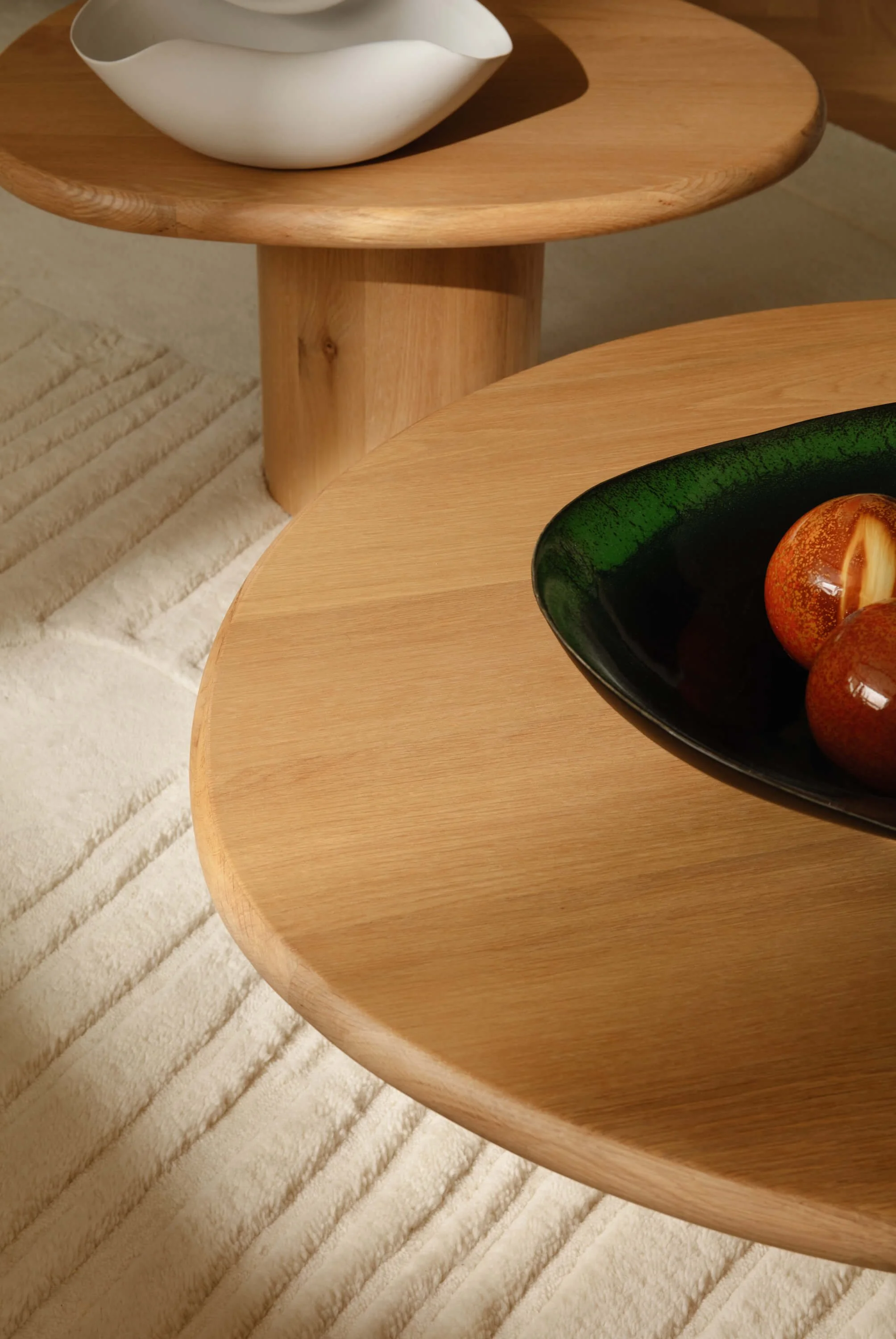 Two round wooden tables with a white ceramic bowl on the upper table and a black ceramic bowl with brown fruits on the lower table, on a striped beige rug.