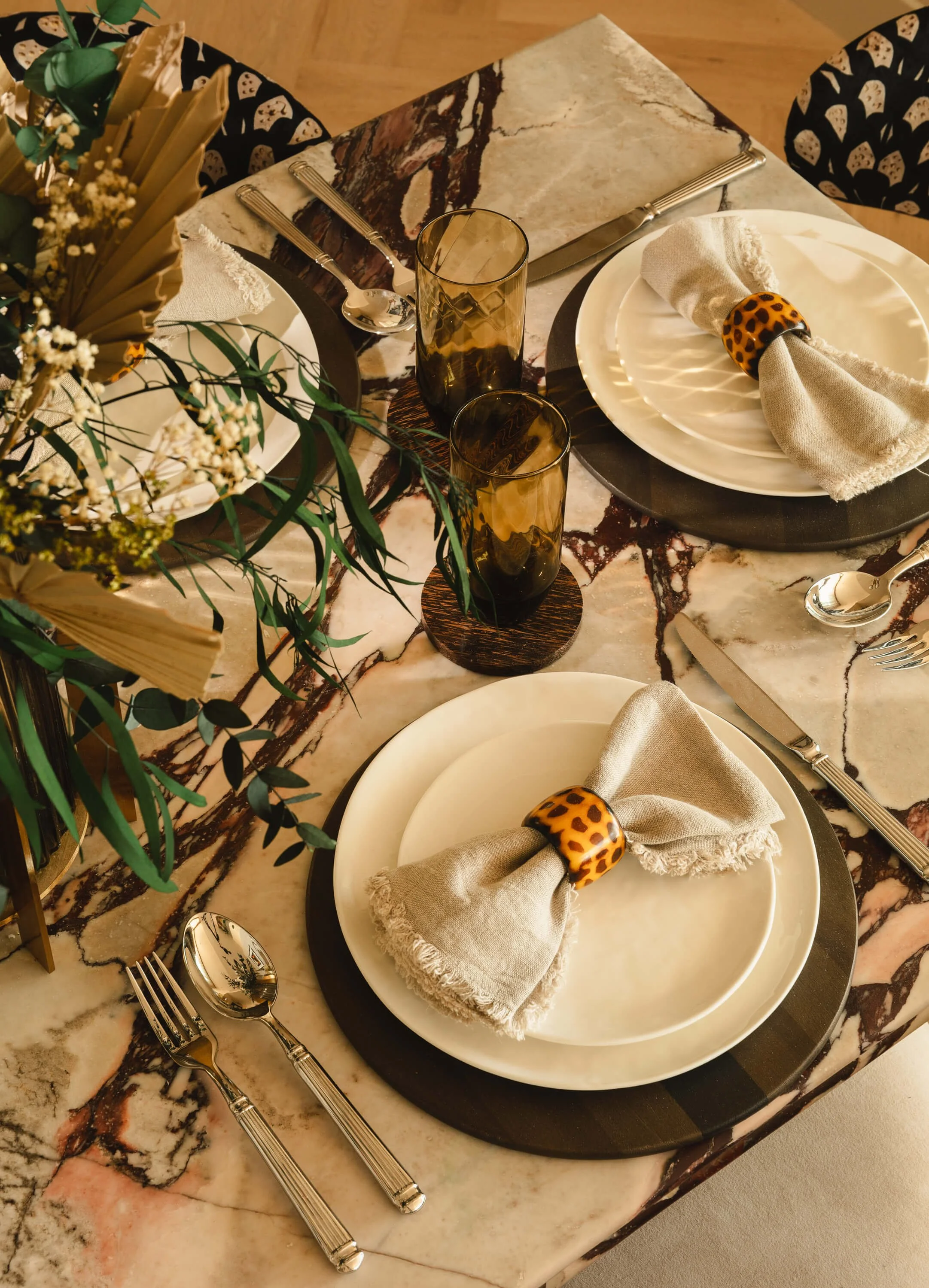 A decorated dining table with white plates, beige cloth napkins secured with leopard print napkin rings, silverware, two amber-colored glasses, and a floral centerpiece.