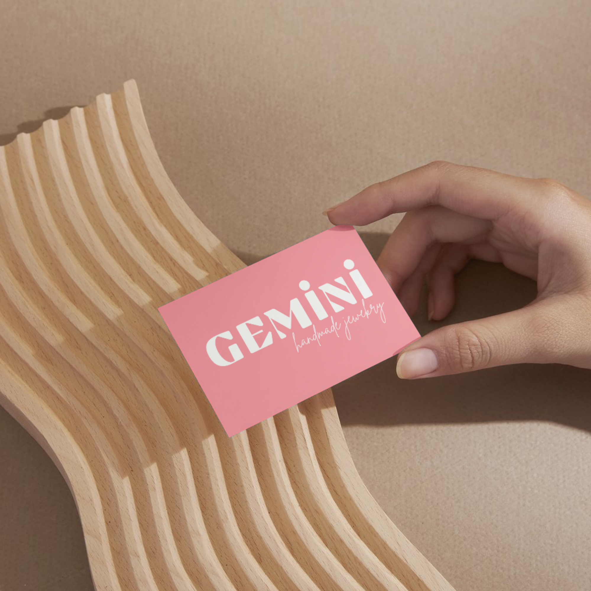 A hand holding a pink business card that reads 'GEMINI handmade jewelry' above a wavy wooden display stand against a beige background.