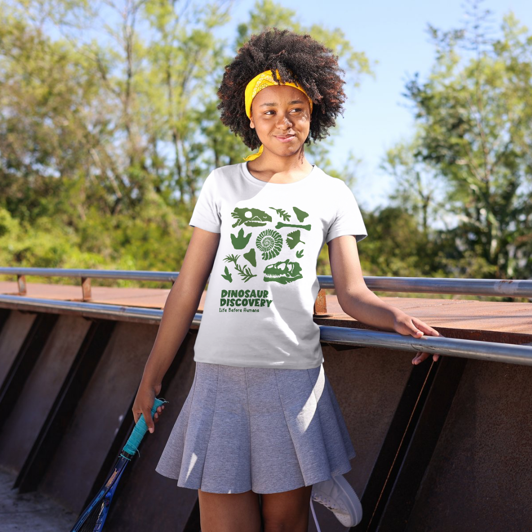 A young girl with curly hair, wearing a yellow headband, white t-shirt with dinosaur silhouettes and the words "Dinosaur Discovery Life Before Humans," and a gray pleated skirt, stands outdoors on a sunny day, holding a tennis racket, with trees and 