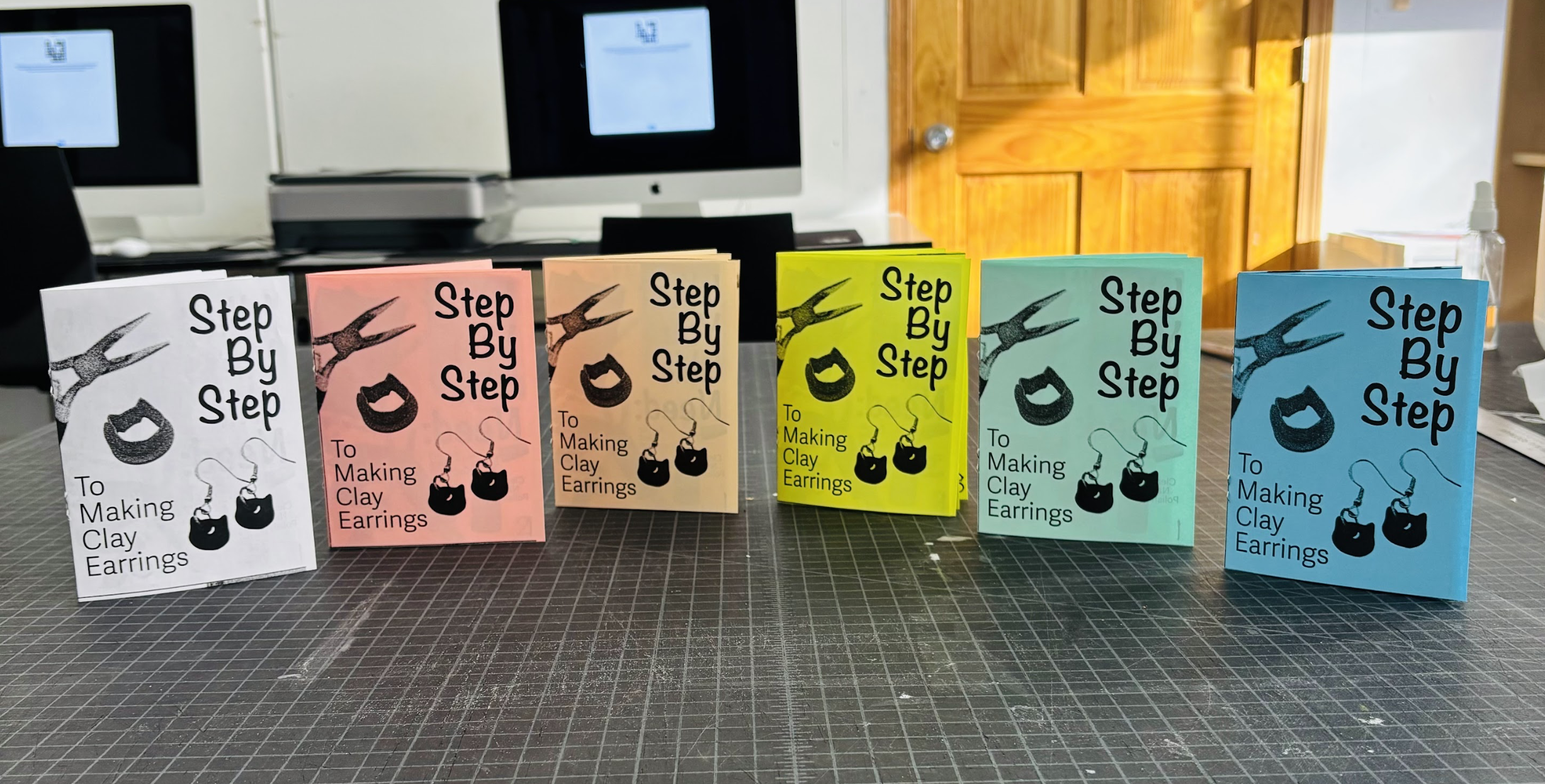 Colorful booklets titled 'Step By Step To Making Clay Earrings' arranged in a row on a grid-patterned surface, with a computer and a wooden door in the background.