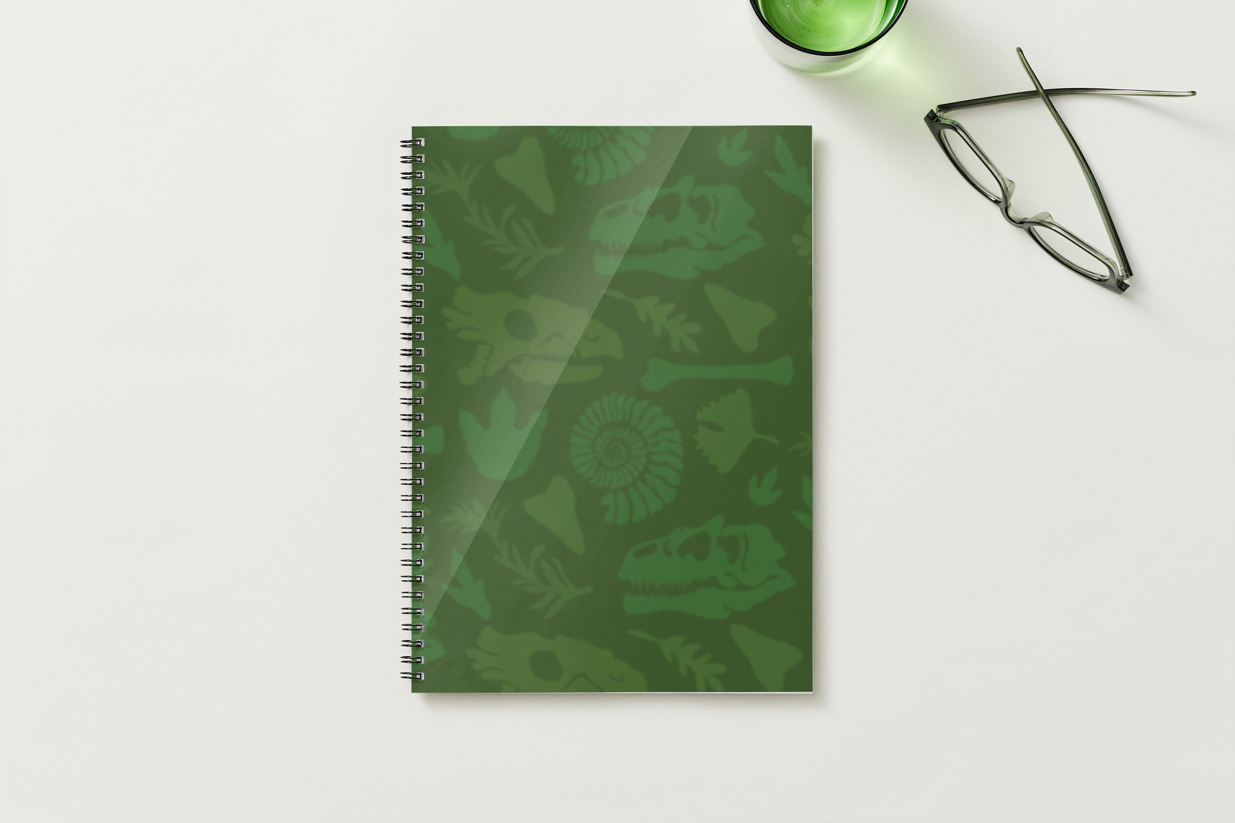 A white desk with a green spiral notebook, eyeglasses, and a green glass of beverage.