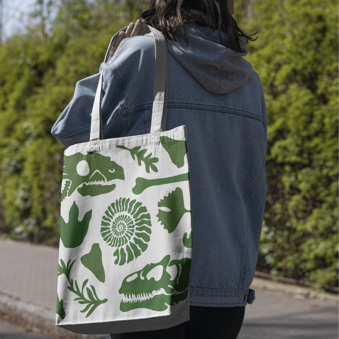 A person in a blue denim jacket with a gray hood carrying a large white tote bag with green dinosaur and shell motifs, walking outdoors on a sunny day with trees in the background.