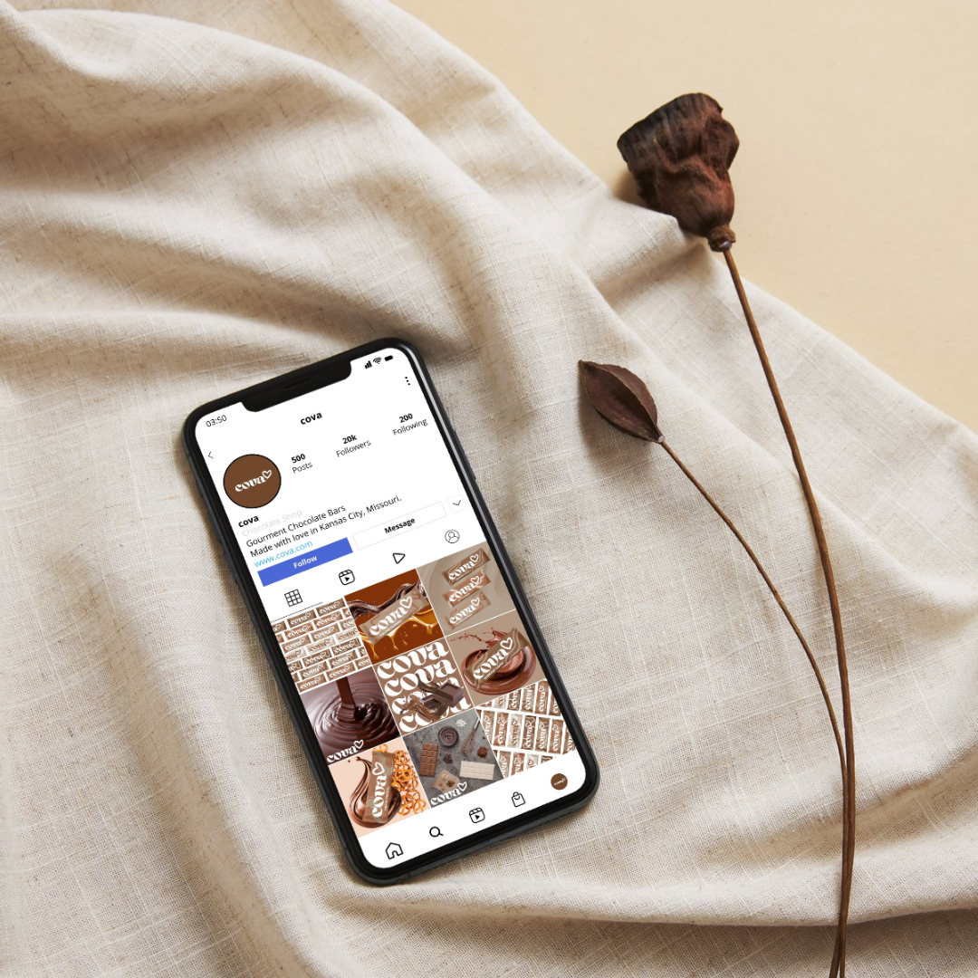 Smartphone displaying an Instagram profile page for 'cova', a chocolate brand, on a beige fabric surface with two dried plants beside it.