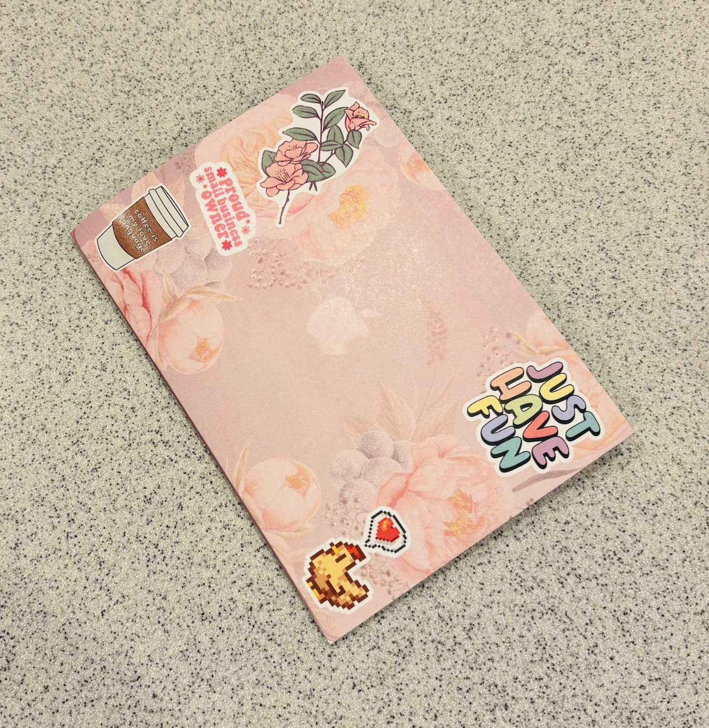 Light pink decorative sheet of paper with stickers of a coffee cup, a flower, and pixel art of a hand with a heart, on a speckled gray surface.